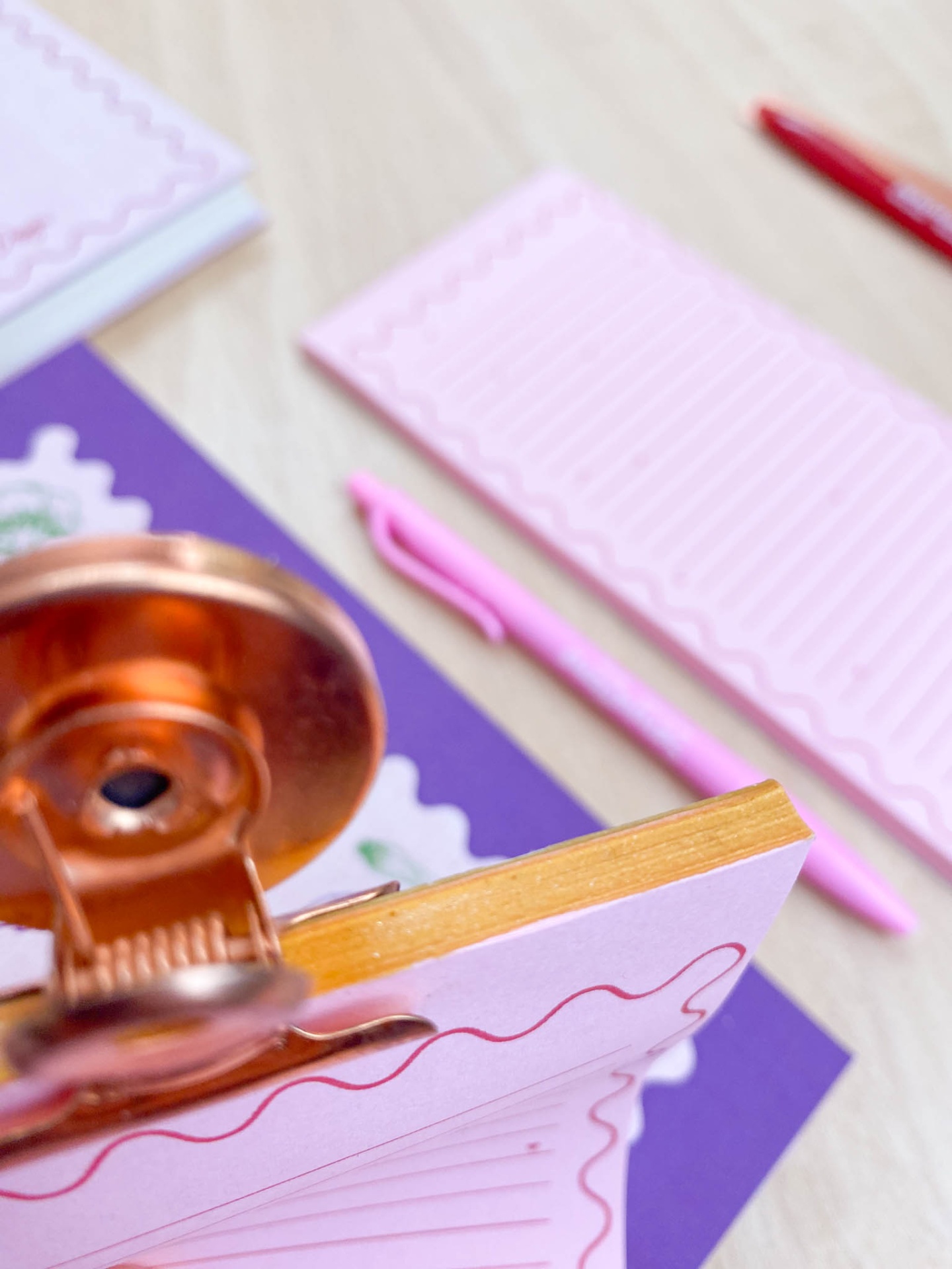 Blocos de notas cor-de-rosa com linhas onduladas e clipes dourados, canetas cor-de-rosa, e fundo lilás sobre uma mesa de madeira clara.