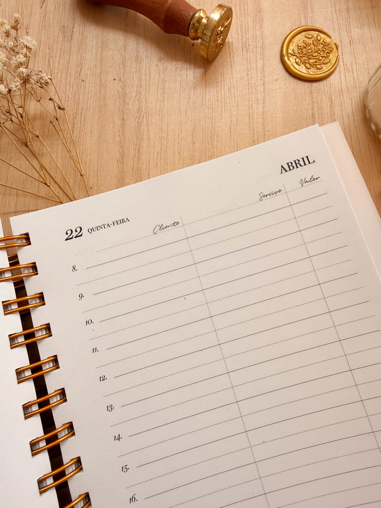 Página de agenda datada 22 Abril com colunas para Cliente, Serviço e Valor sobre mesa de madeira com flores secas e selo de cera