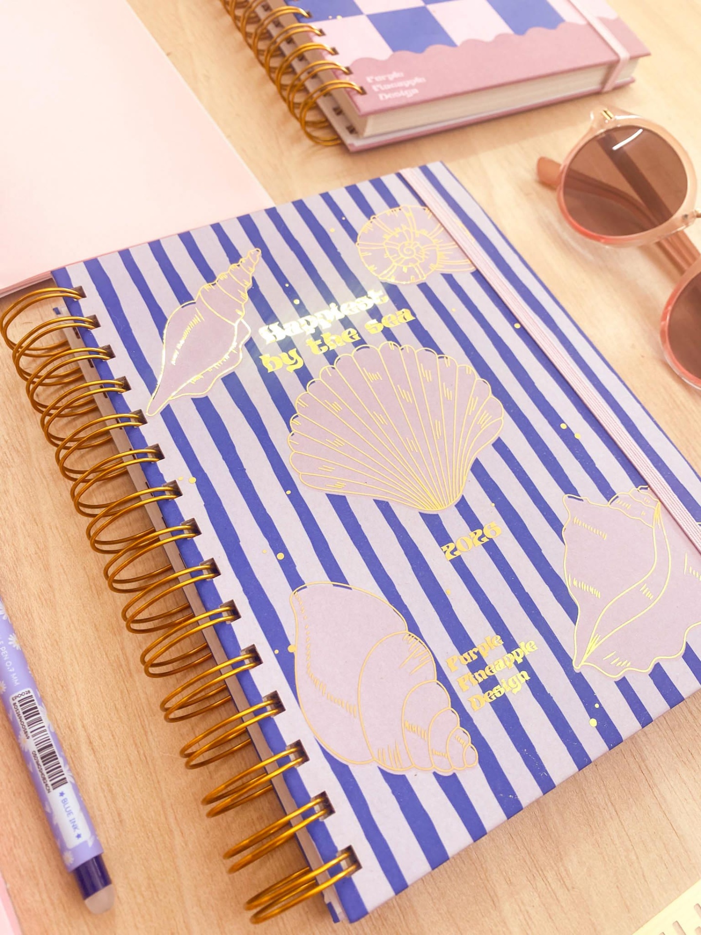 Caderno de capa dura riscada azul e branca com conchas douradas e texto, encadernação espiral dourada, sobre mesa de madeira clara com caneta, outro caderno e óculos de sol