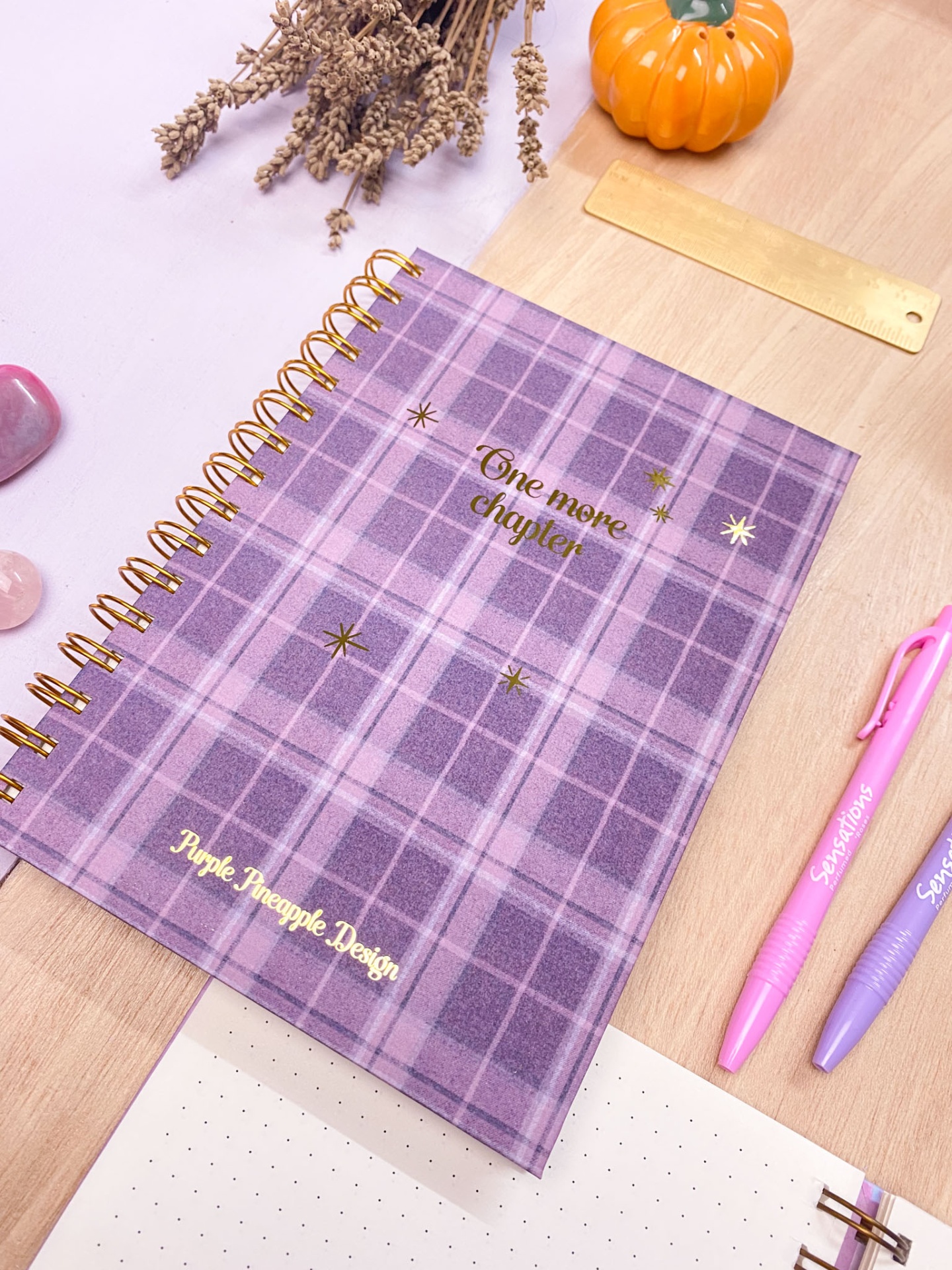 Caderno xadrez lilás com texto dourado e espiral dourada sobre mesa com acessórios de escrita e decoração