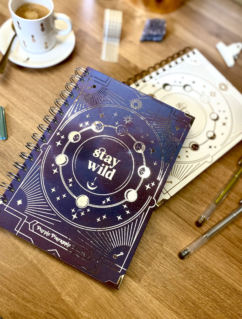 Dois cadernos espiralados com tema lunar e canecas de café numa mesa de madeira.