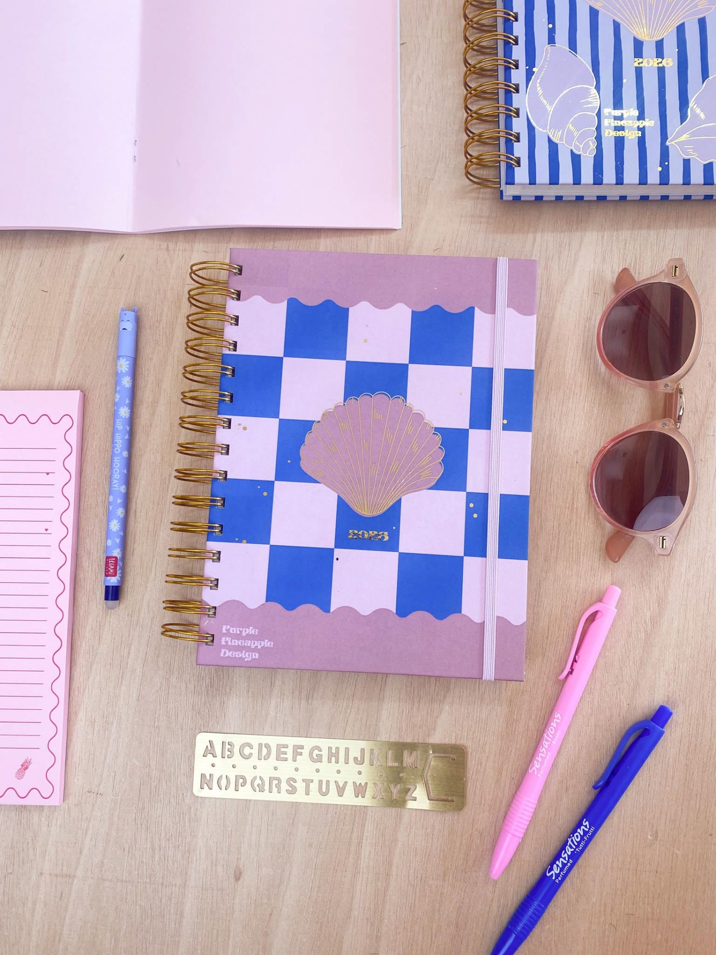 Caderno espiral azul e lilás com canetas, etiquetas, régua e óculos de sol