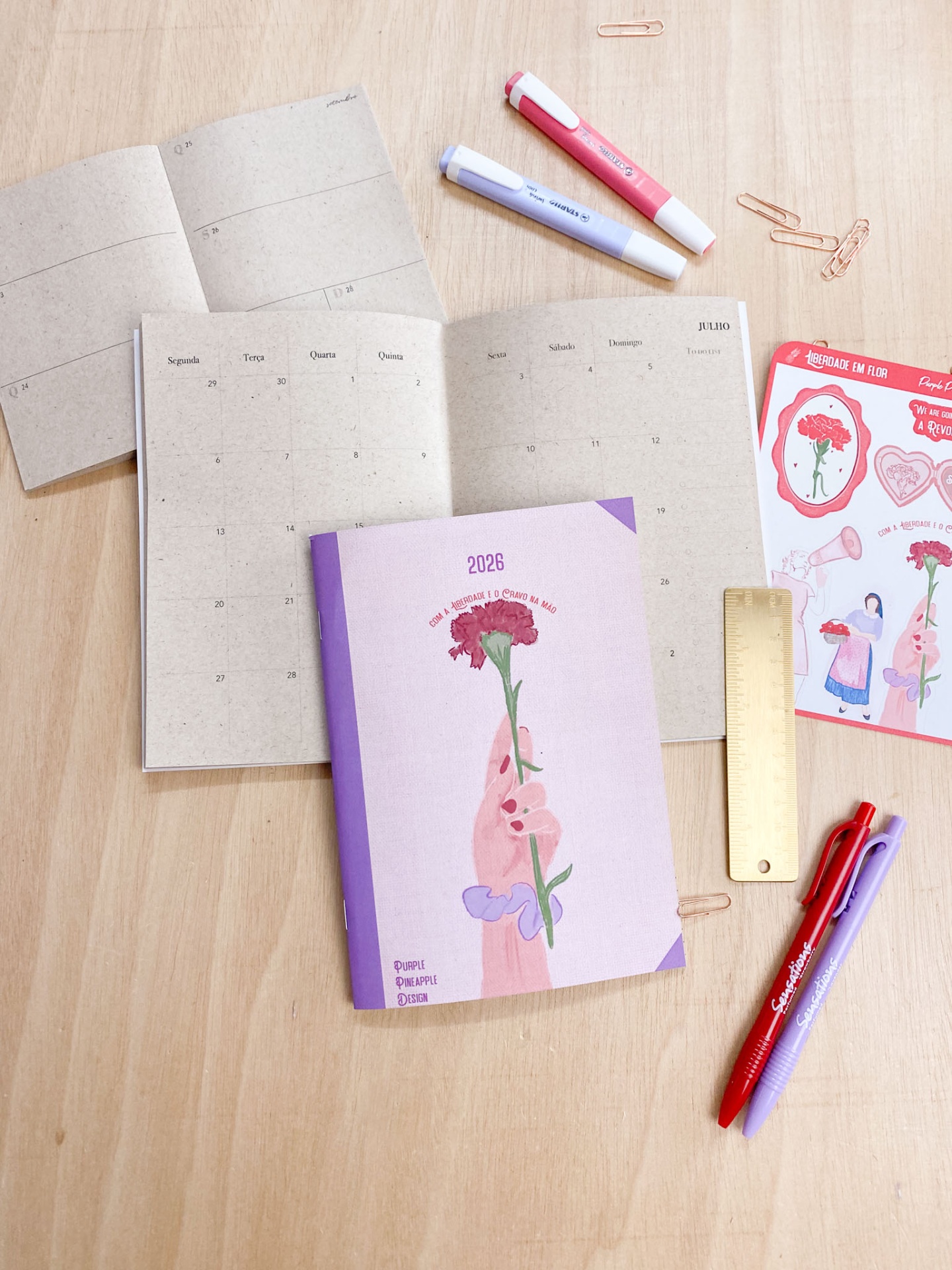 Agenda ilustrada com canetas, régua dourada, clips e autocolantes numa mesa de madeira clara