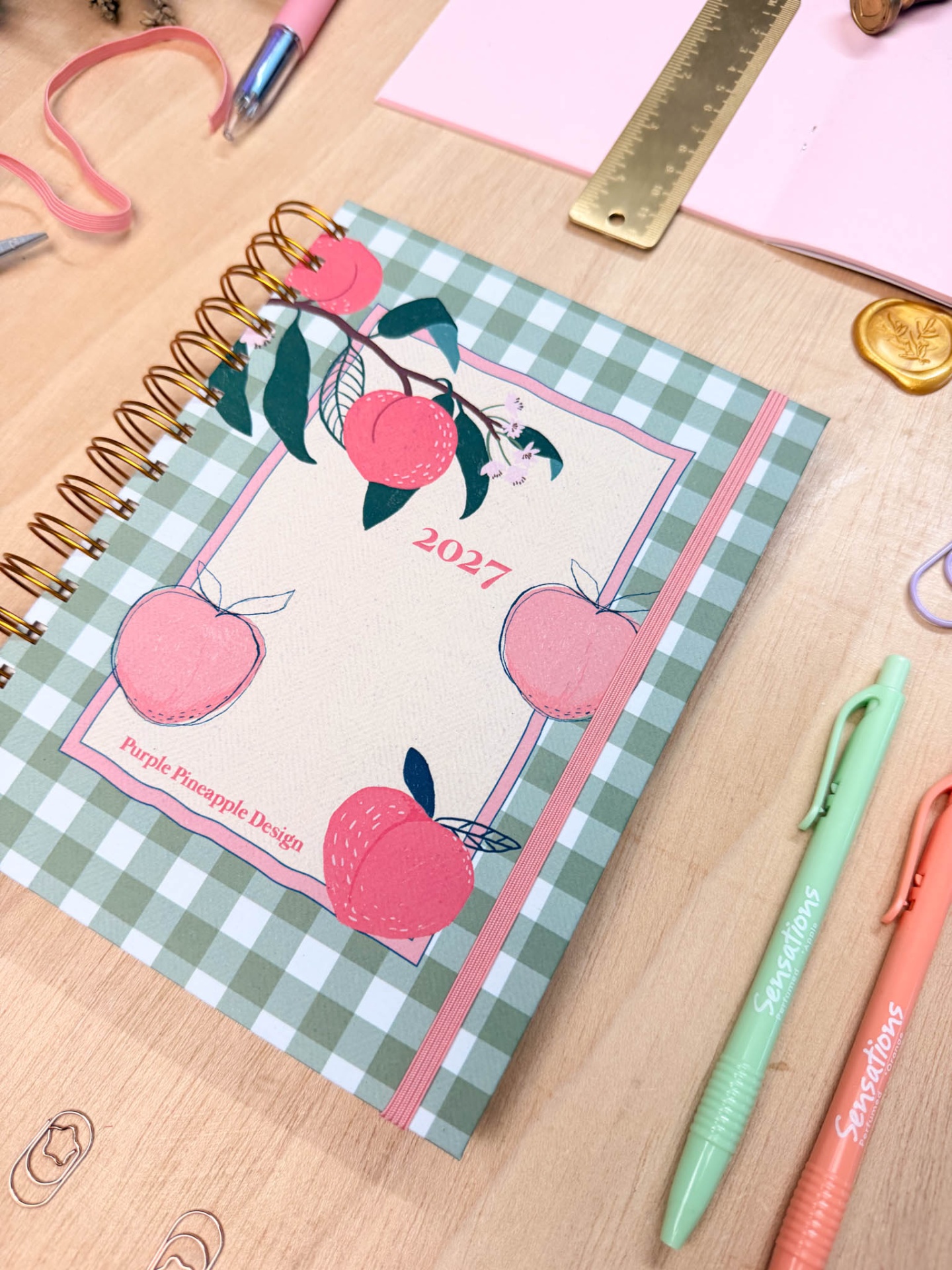 Caderno espiral decorado com padrão xadrez e ilustração de pêssegos cor-de-rosa numa mesa com canetas e régua