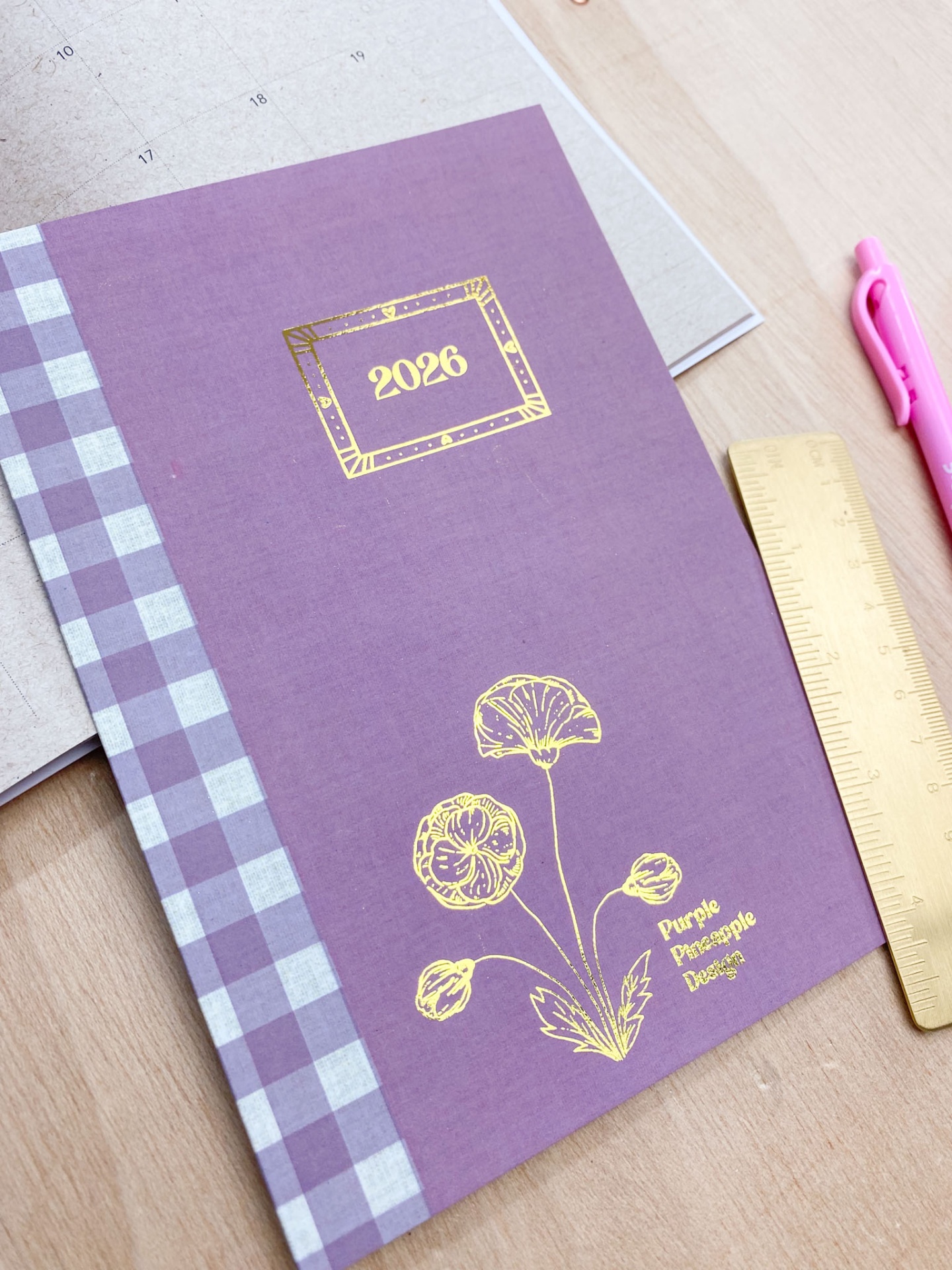 Agenda ou caderno roxo com detalhes florais dourados e texto '2026' em ambiente de mesa com régua e caneta