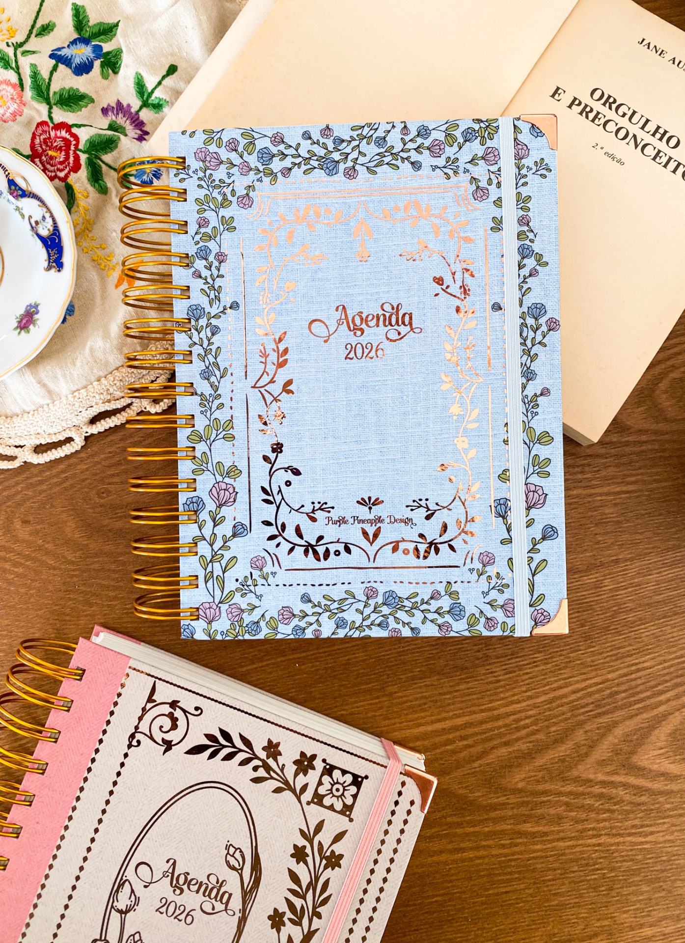 Duas agendas 2026 com design floral e detalhes dourados sobre mesa de madeira, ao lado de um livro e uma chávena