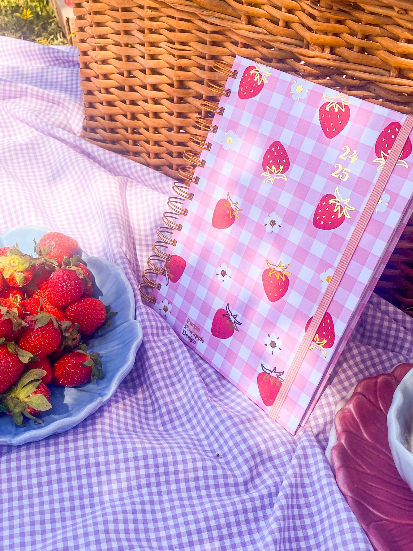 Diário rosa com morangos estampados sobre pano lilás xadrez ao lado de cesta de vime e prato azul com morangos