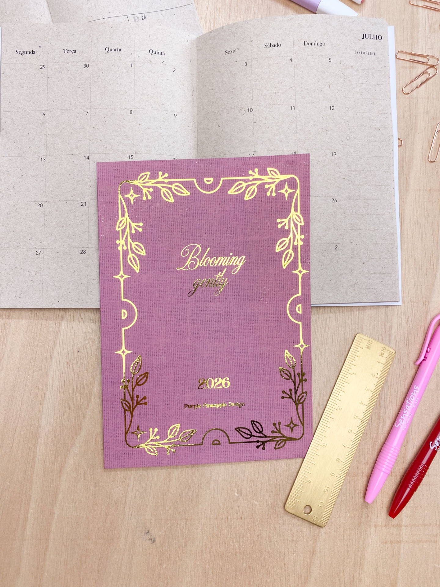 Agenda rosa 2026 decorada com dourado sobre mesa de madeira com canetas e régua