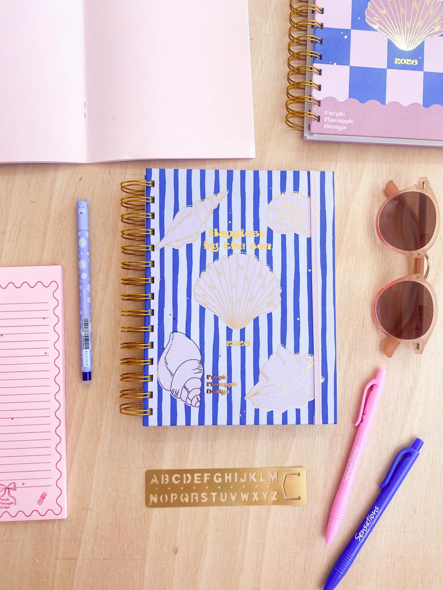 Caderno azul às riscas com conchas, canetas, régua de alfabeto e óculos de sol sobre mesa de madeira