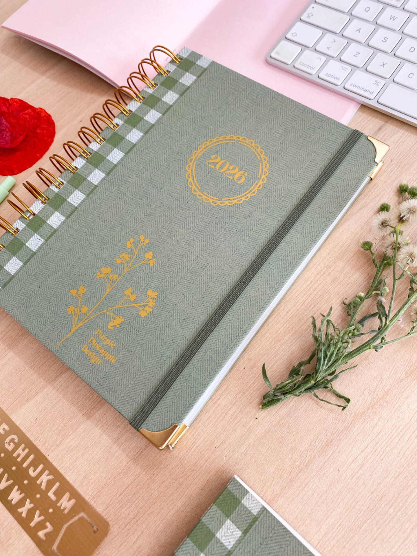 Caderno verde com elástico e espiral dourada sobre mesa de madeira com teclado e flores