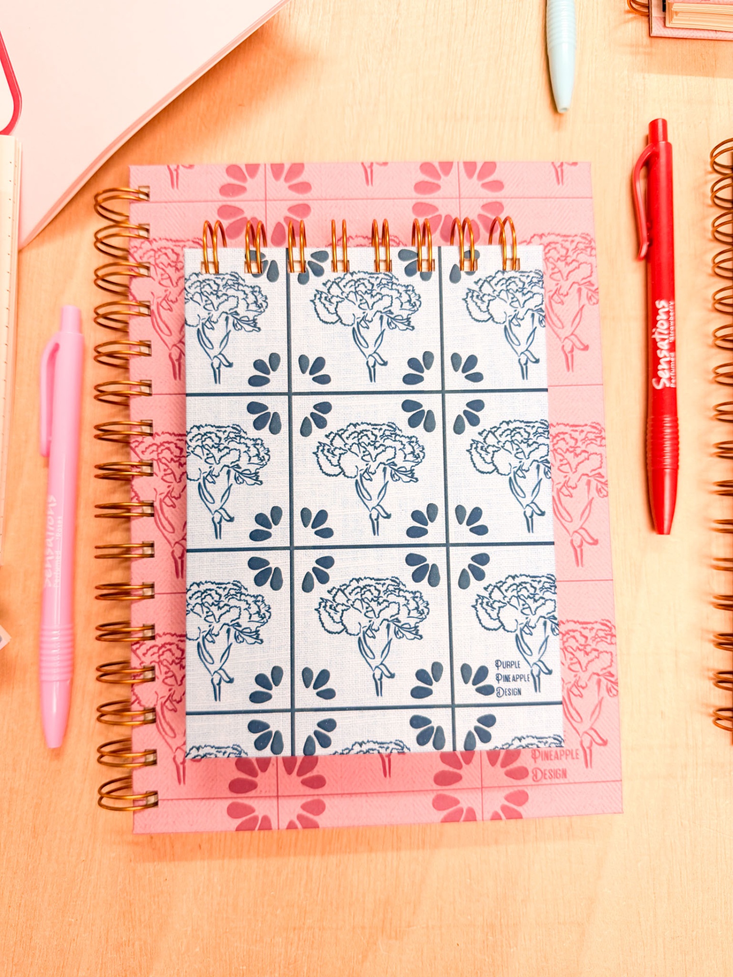 Três cadernos espiralados com padrões florais e duas esferográficas em cima de uma mesa de madeira