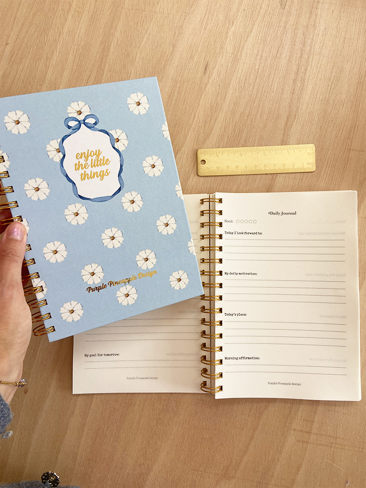 Dois cadernos espirais azuis com flores, um fechado com texto e outro aberto mostrando páginas internas, sobre superfície de madeira clara com régua amarela ao lado.