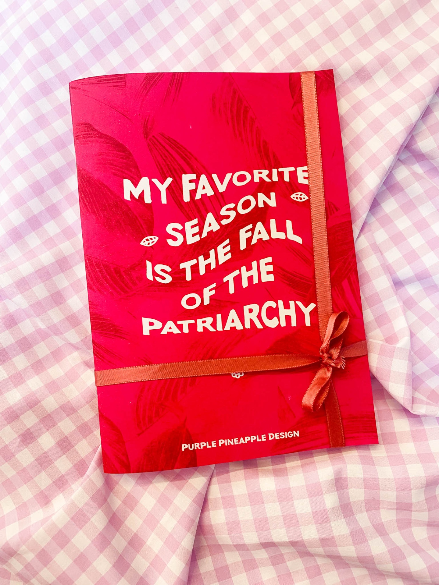 Caderno vermelho com texto branco e fita cor de rosa sobre tecido xadrez rosa e branco