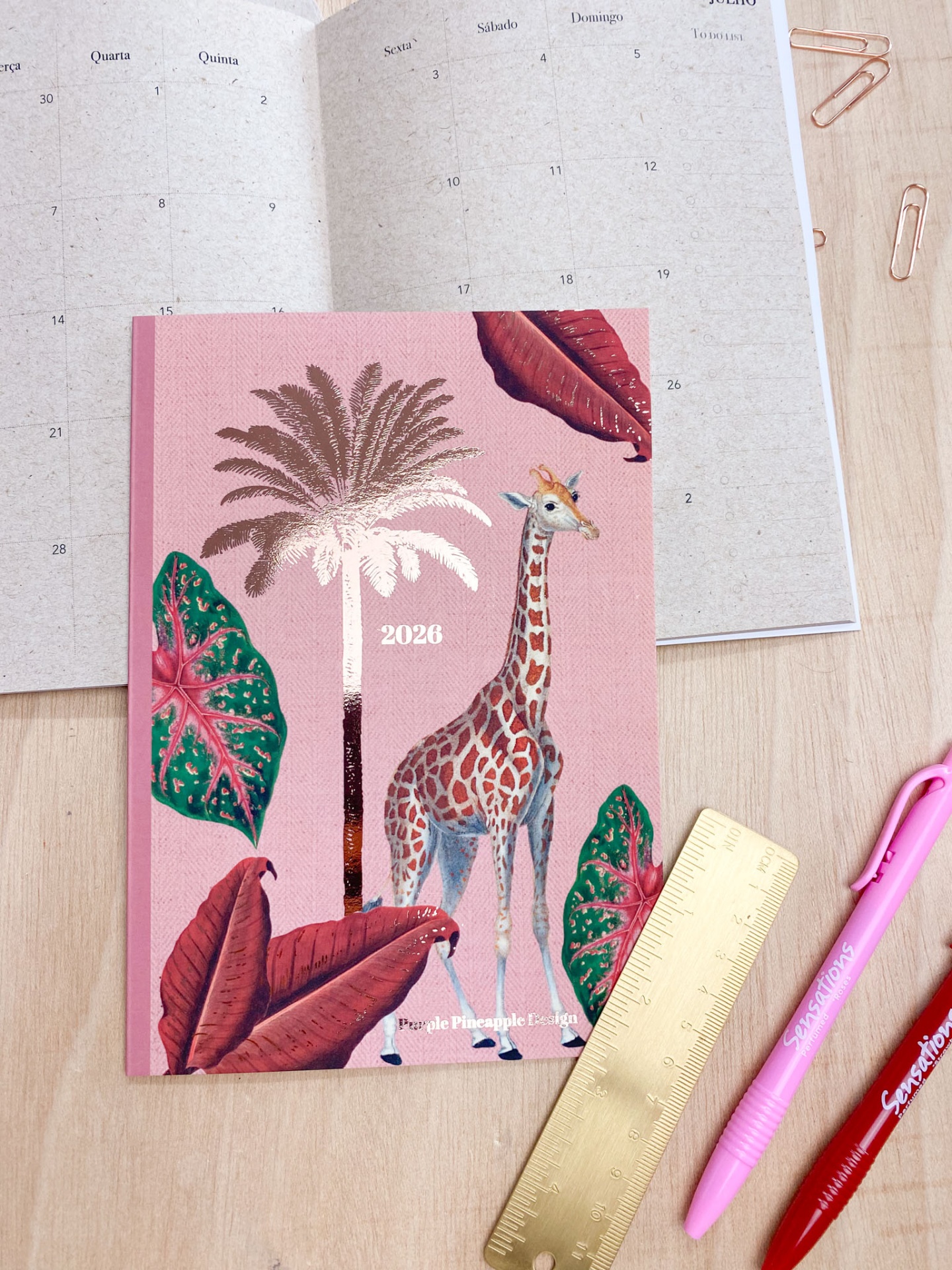 Agenda 2026 com ilustração de girafa e folhas coloridas sobre mesa com régua e canetas.