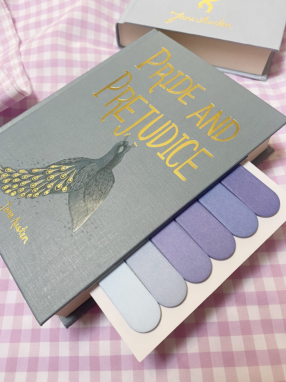 Caixa cinzenta com capa de livro Pride and Prejudice e amostras de tecidos em tons de azul e roxo sobre tecido rosa xadrez
