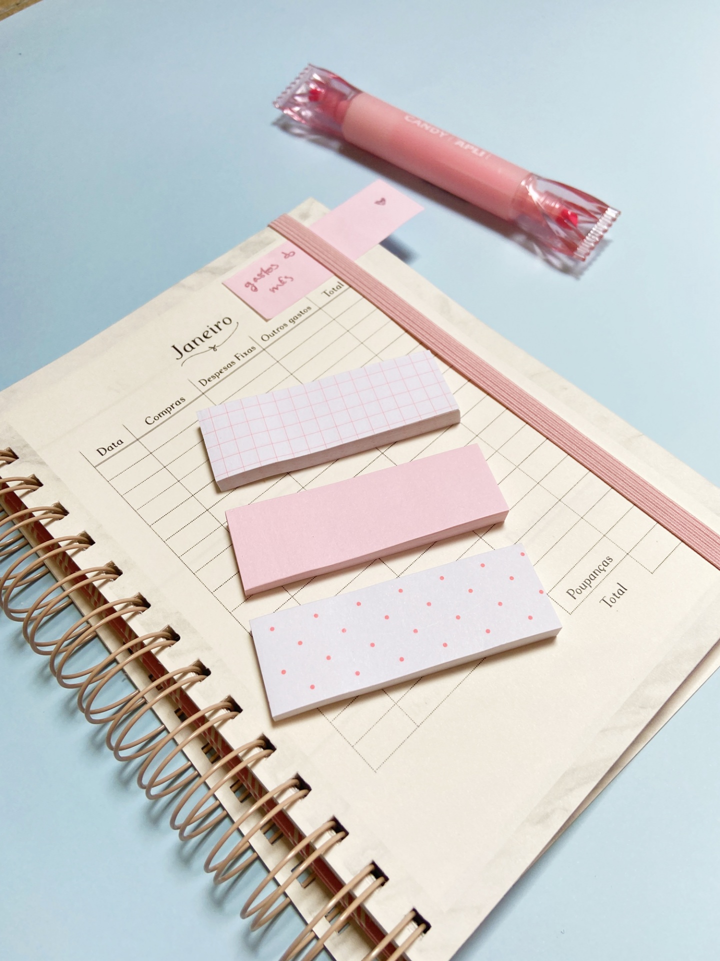 Caderno de argolas com folhas creme e notas autocolantes em tons de rosa e branco sobre fundo azul claro.