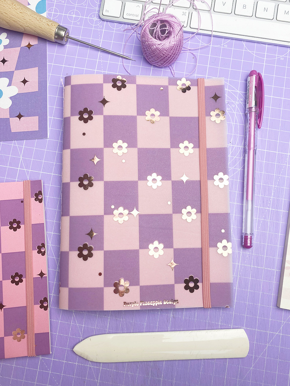 Caderno rosa e lilás com padrão xadrez e flores em relevo numa mesa lilás com acessórios de artesanato