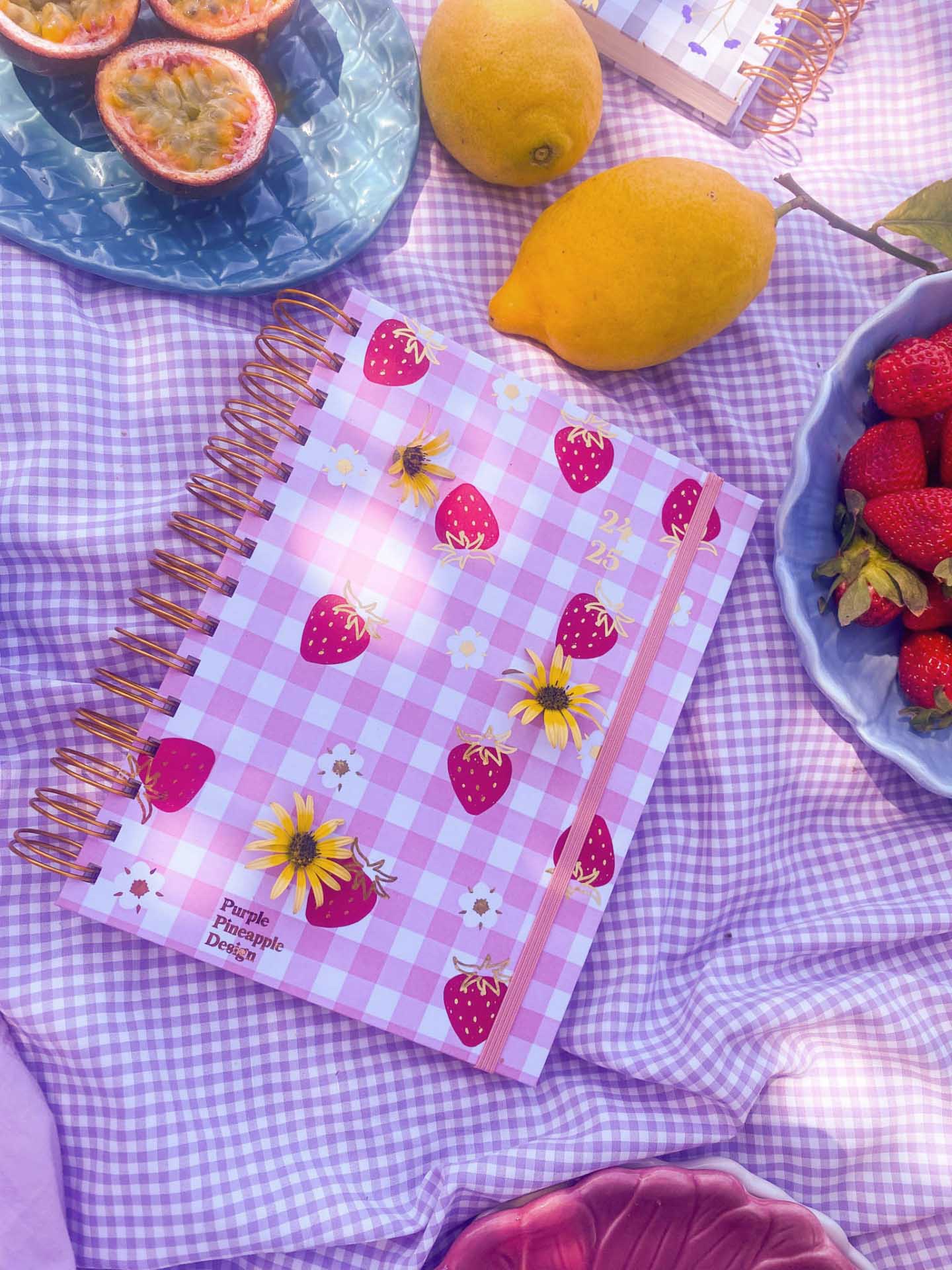 Caderno espiral roxo com morangos e flores, rodeado por limões, morangos e maracujás sobre tecido roxo aos quadrados.