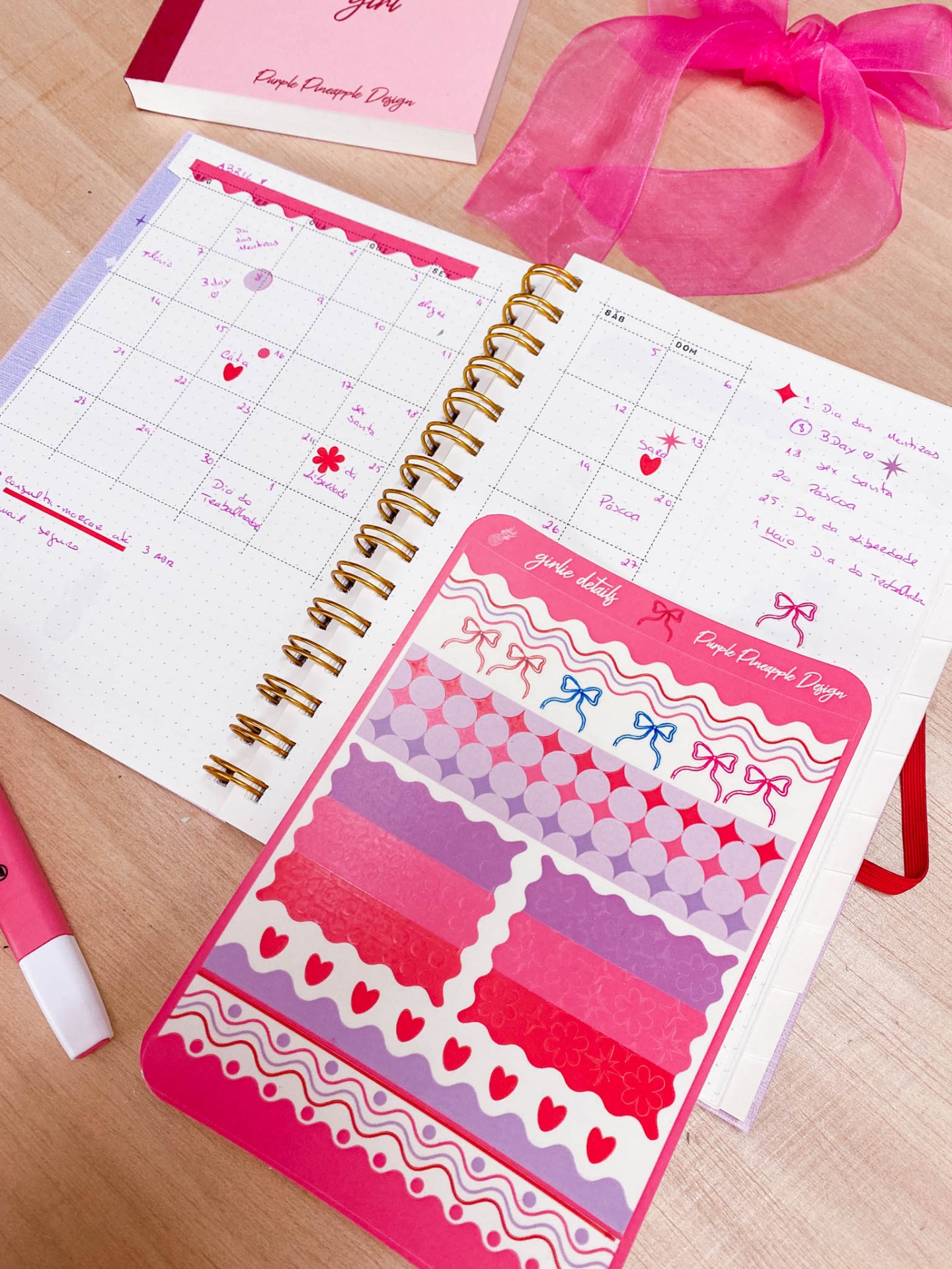 Caderno de planejamento mensal aberto com anotações e sticker de fitas coloridas em rosa, vermelho e lilás
