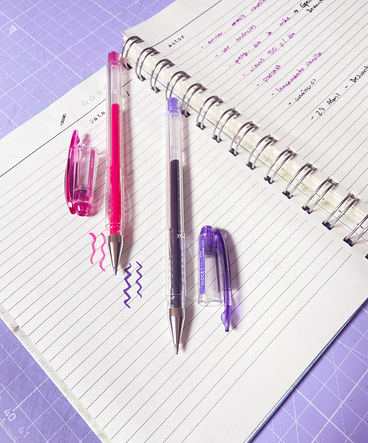 Duas canetas esferográficas transparentes rosa e roxa sobre caderno com escrita colorida