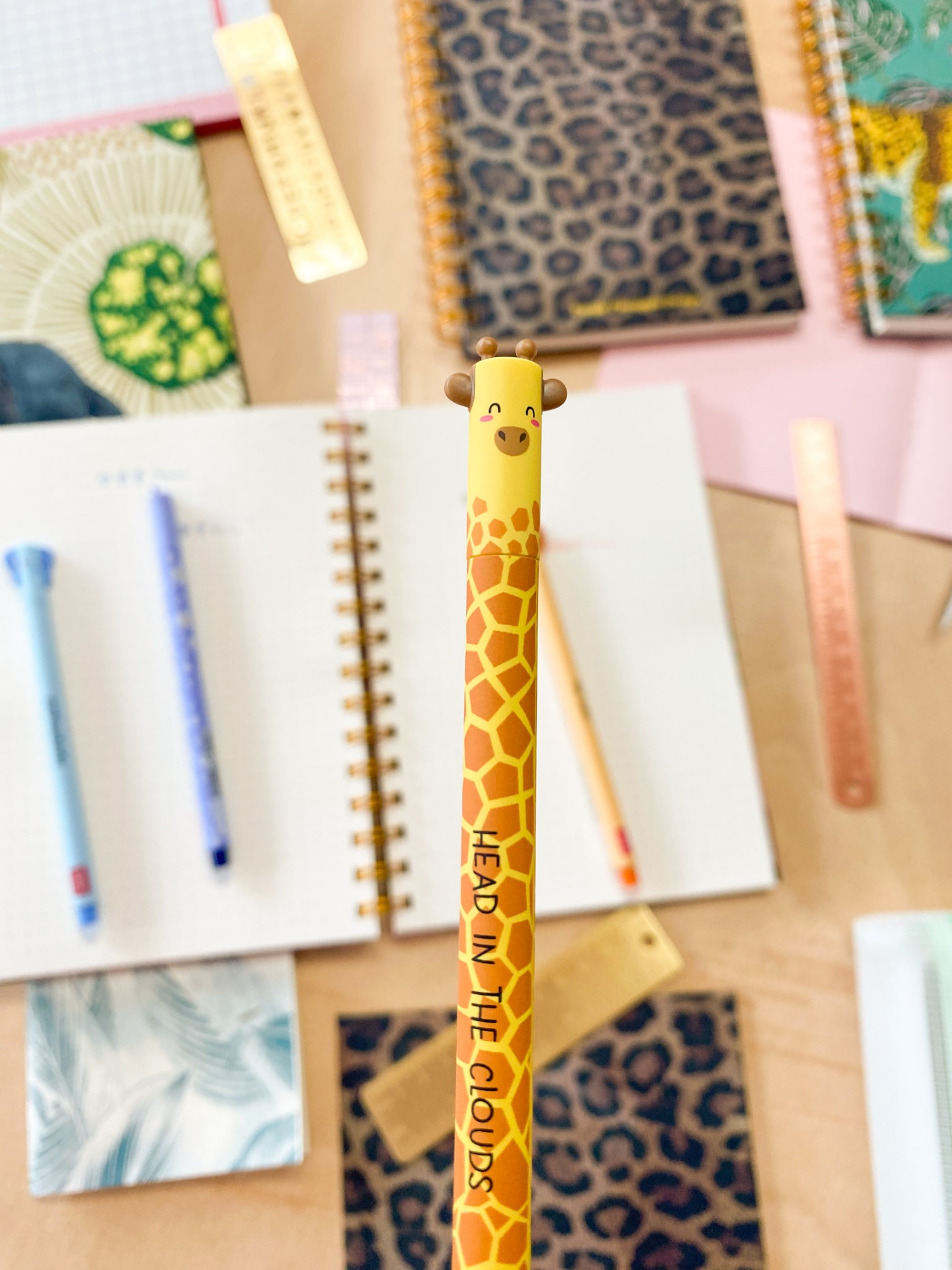 Lápis amarelo com padrão de girafa e texto 'HEAD IN THE CLOUDS' sobre mesa com cadernos e réguas