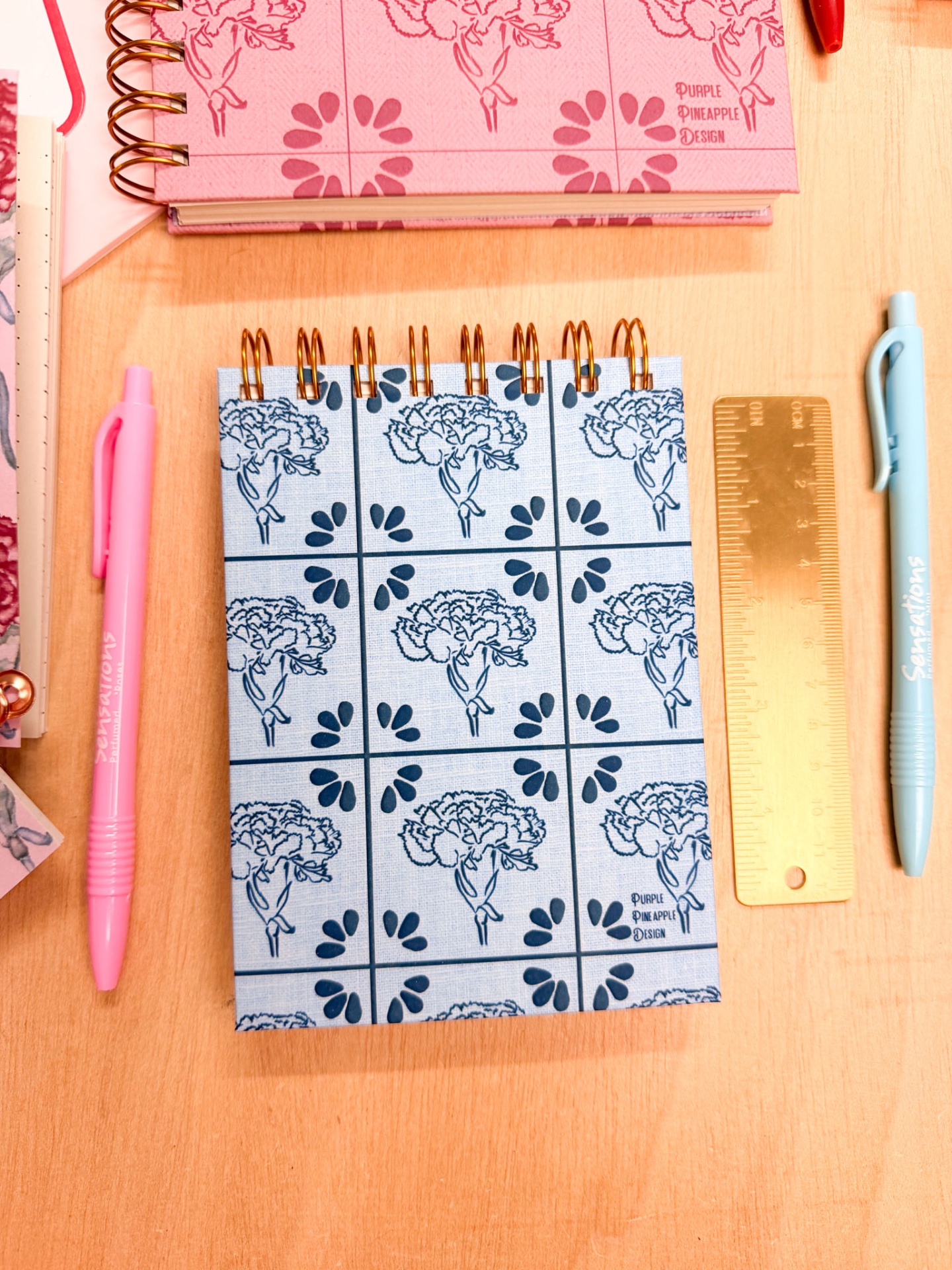 Caderno azul claro com padrão de flores e gotas em espiral dourada, régua dourada e canetas cor-de-rosa e azul-claro