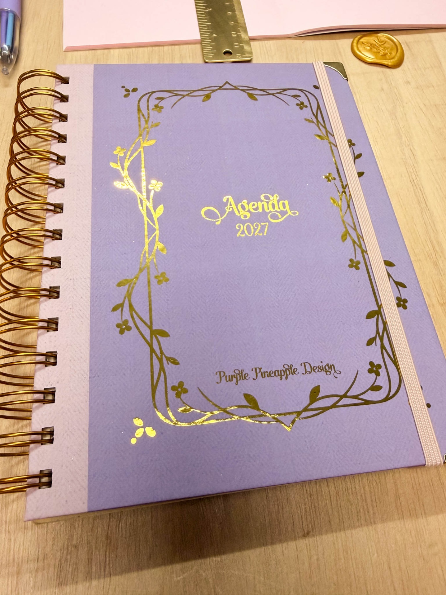 Agenda 2027 lilás com detalhes dourados e espiral dourado sobre mesa de madeira