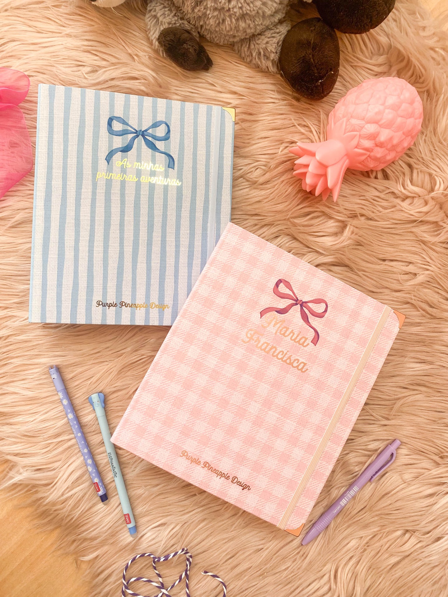 Dois cadernos em tecido azul às riscas e rosa às quadrículas, com texto personalizado, elástico dourado, canetas coloridas, brinquedo em forma de ananás cor-de-rosa e peluche sobre superfície peluda castanha.