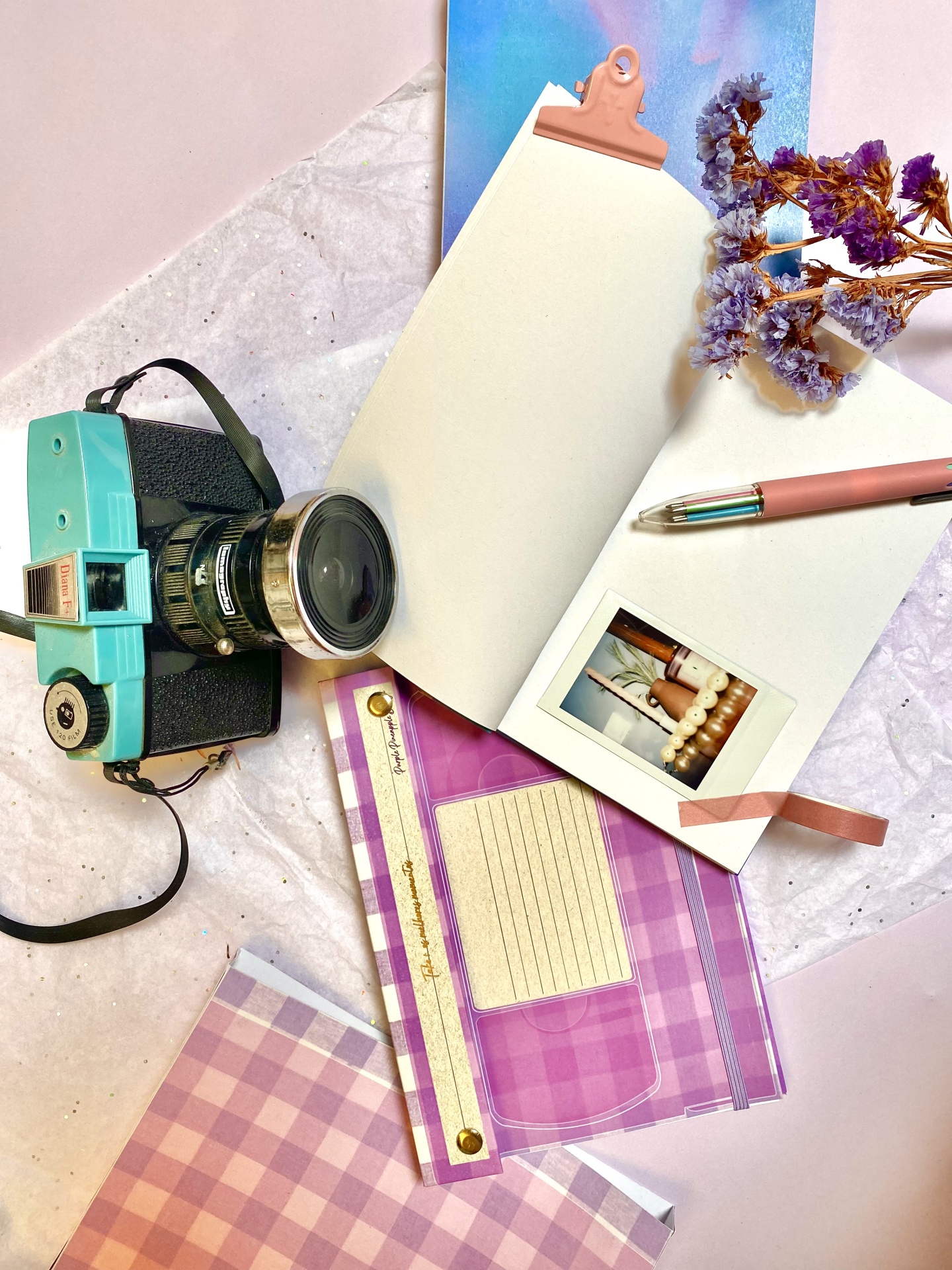Como Criar um Vision Board no Teu Journal Câmara fotográfica retro azul com caderno aberto, caneta rosa, régua, papel xadrez lilás e flores secas roxas