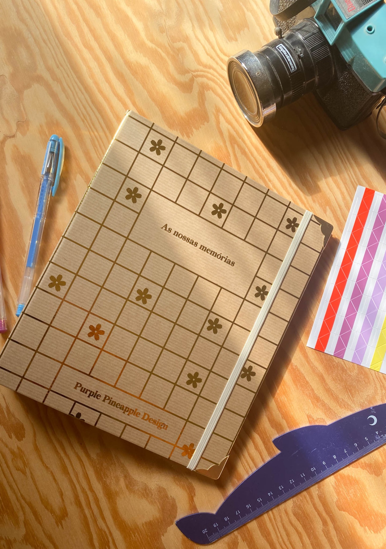 Caderno decorado castanho com texto e padrão floral, com caneta azul, régua curva roxa, câmara e fitas coloridas sobre mesa de madeira