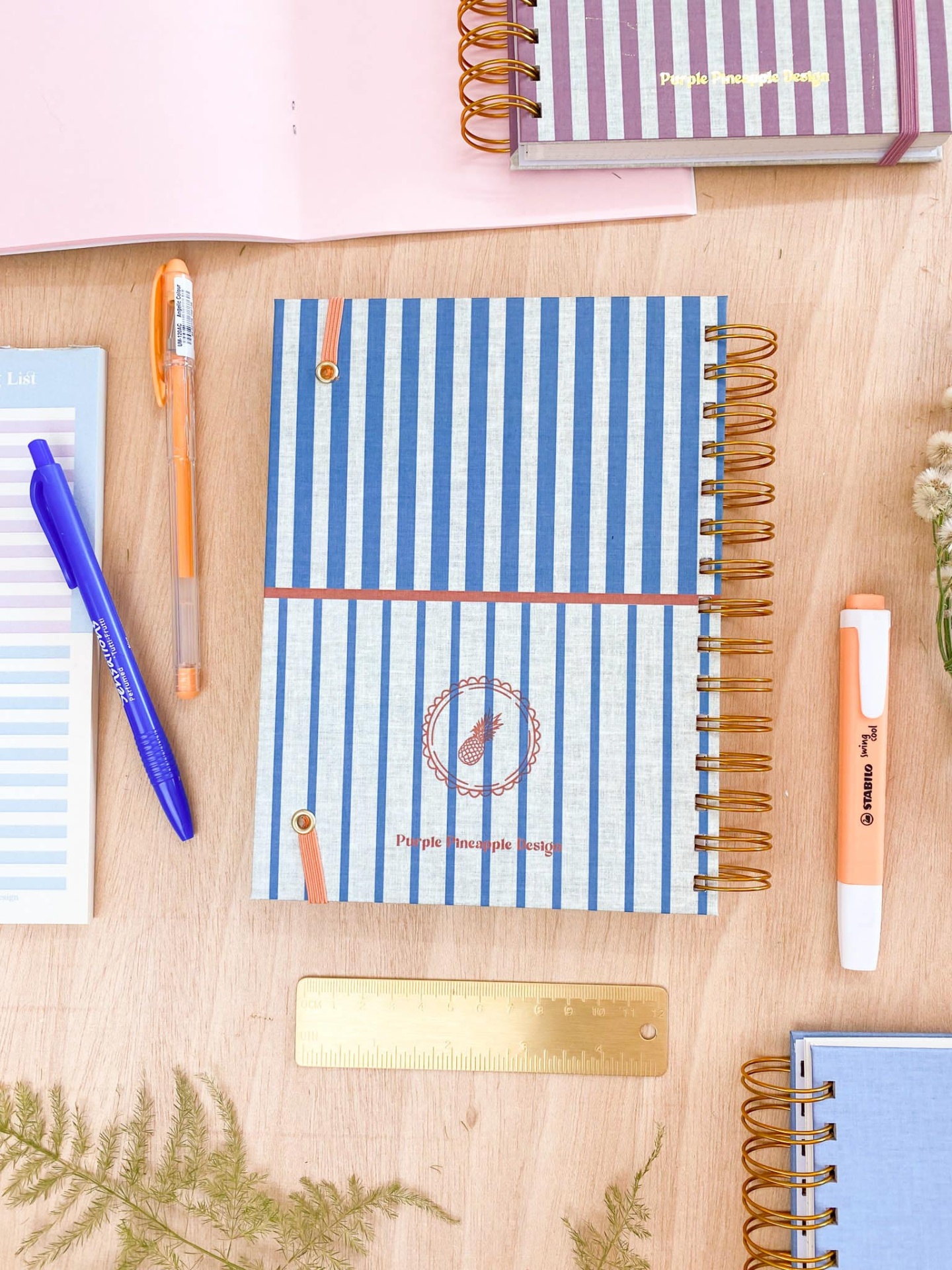 Caderno espiral riscas azuis brancas com canetas e régua sobre mesa de madeira