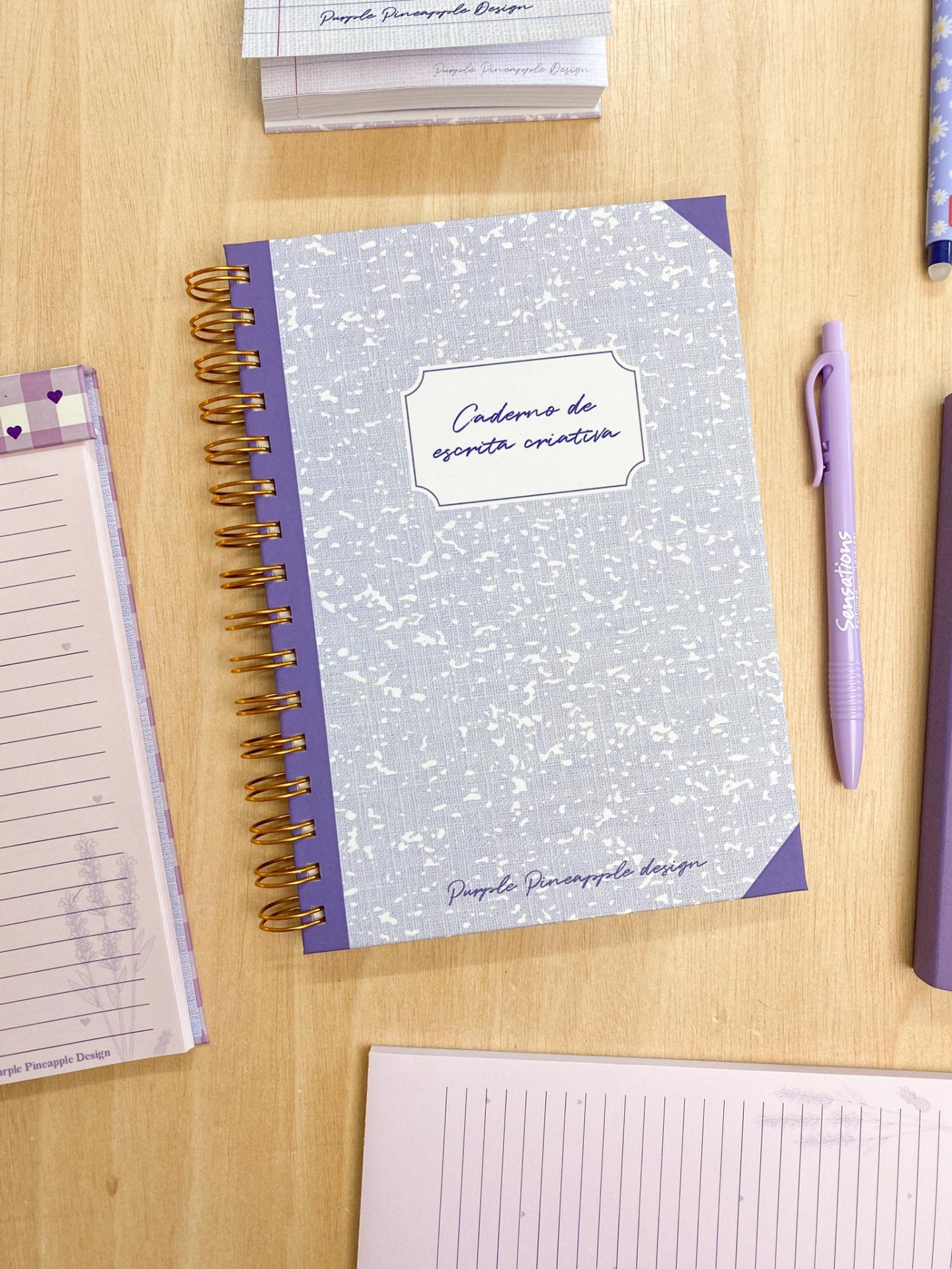 Caderno de escrita criativa com capa cinza e roxa, espiral dourada e caneta roxa sobre mesa de madeira.