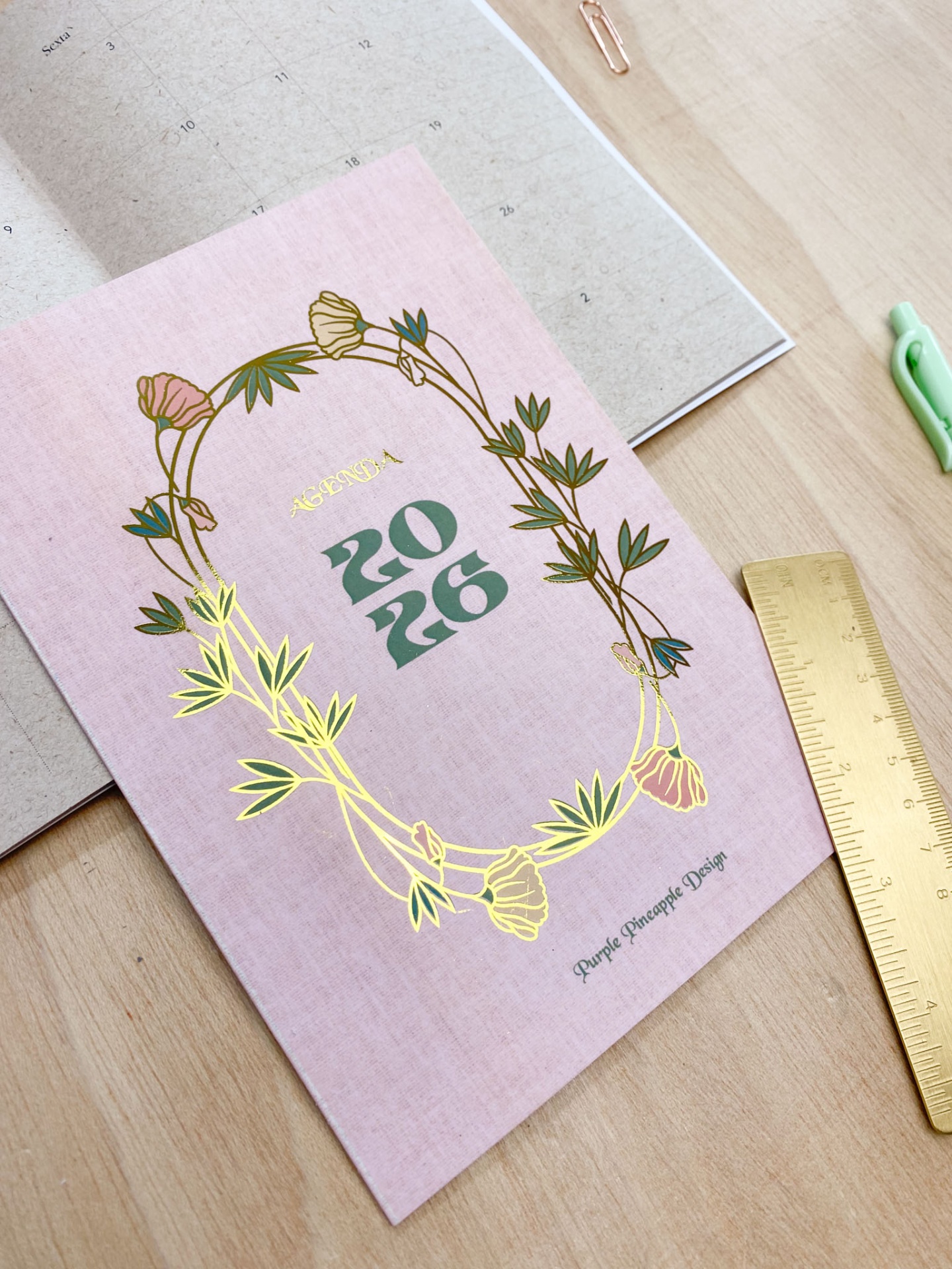 Agenda 2026 com capa rosa e detalhes dourados, régua dourada e caneta verde sobre mesa de madeira clara