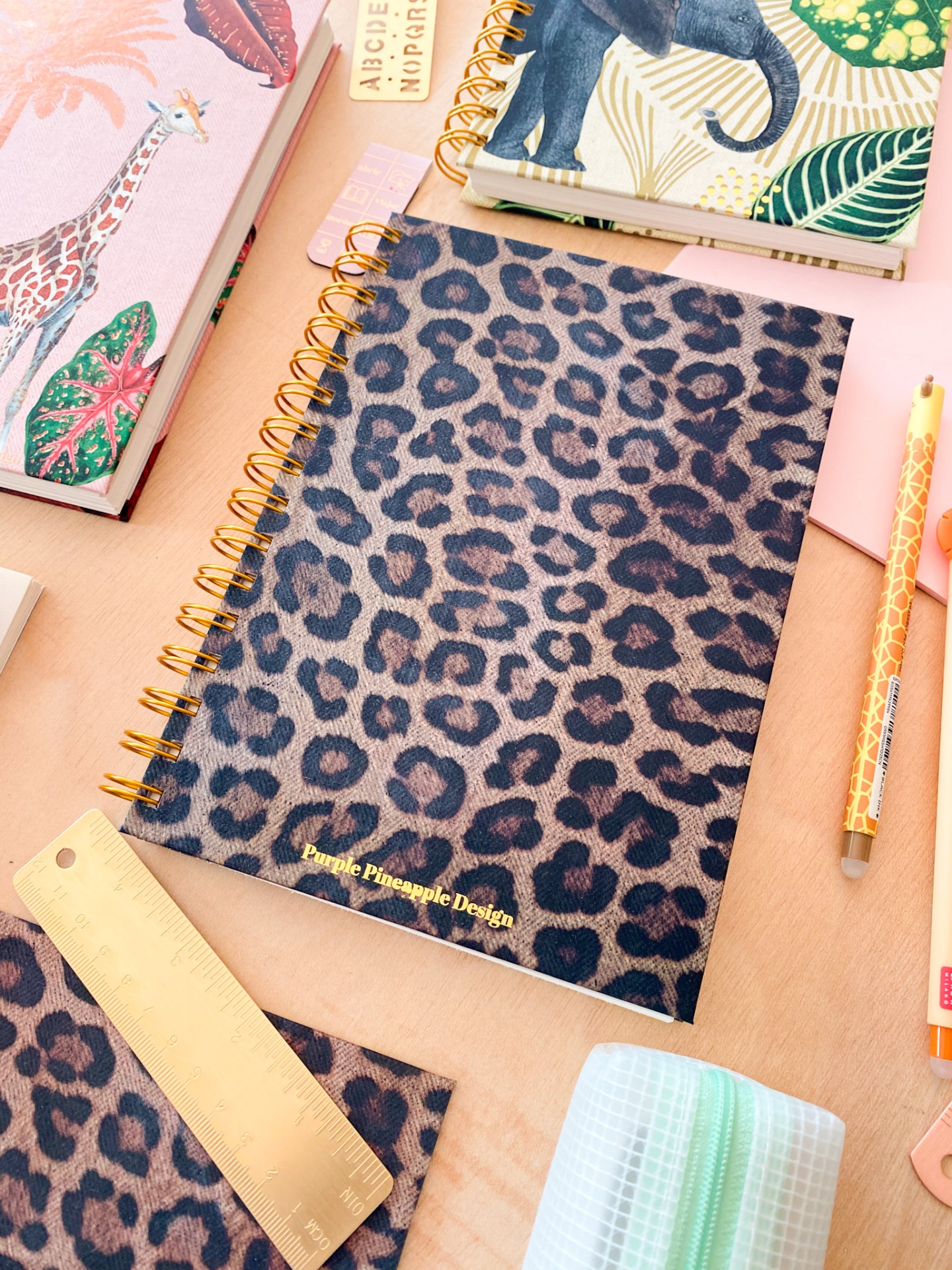 Caderno com capa animal print e espiral dourada ao lado de régua, lápis e outros cadernos coloridos