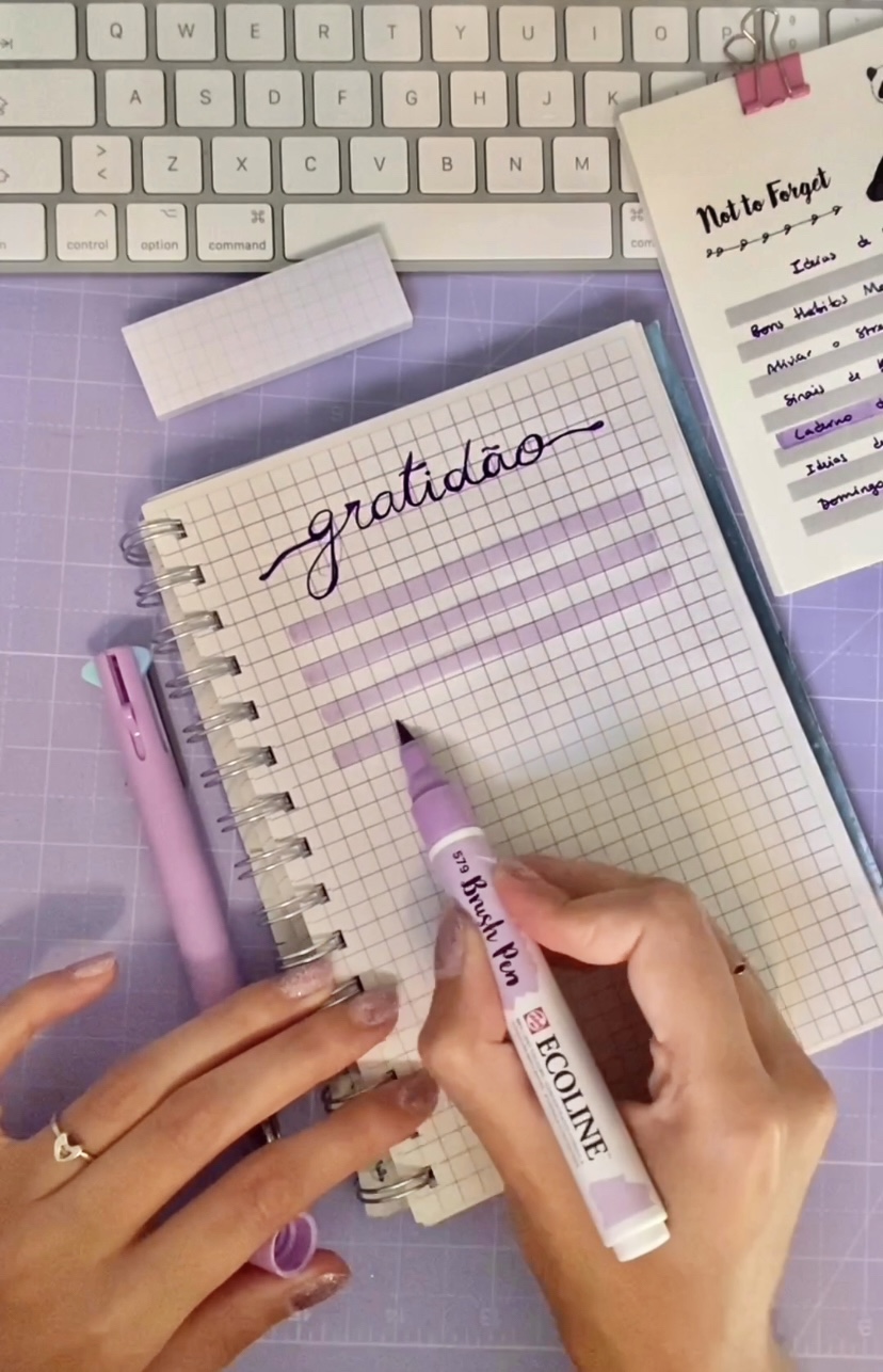 Mão escrevendo 'gratidão' num caderno quadriculado com caneta pincel lilás junto a teclado e outros materiais de escritório