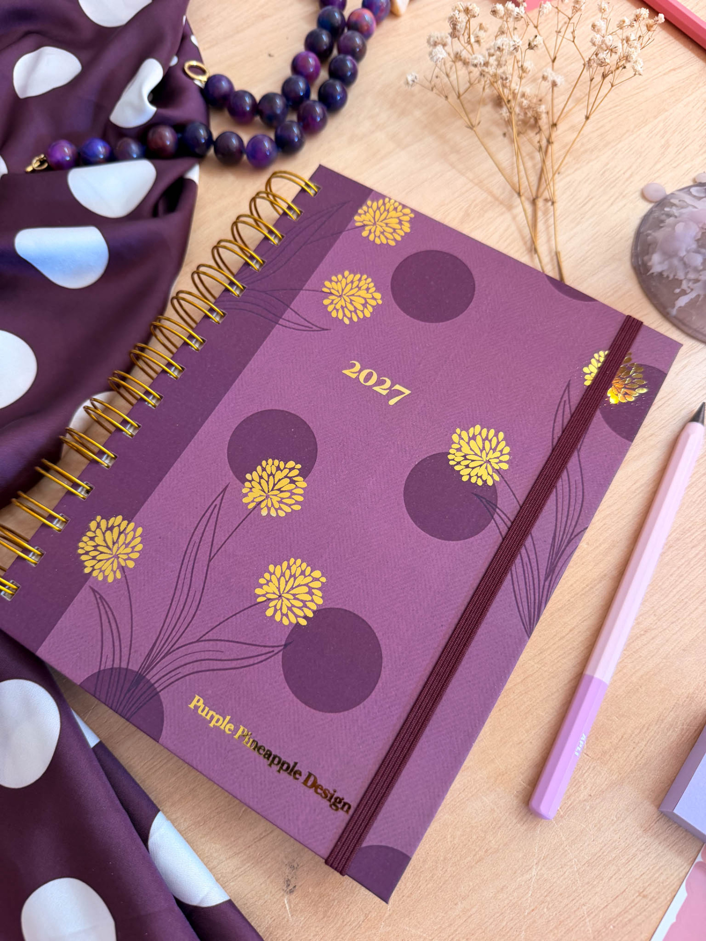 Agenda 2027 com capa roxa floral e elástico sobre mesa com acessórios