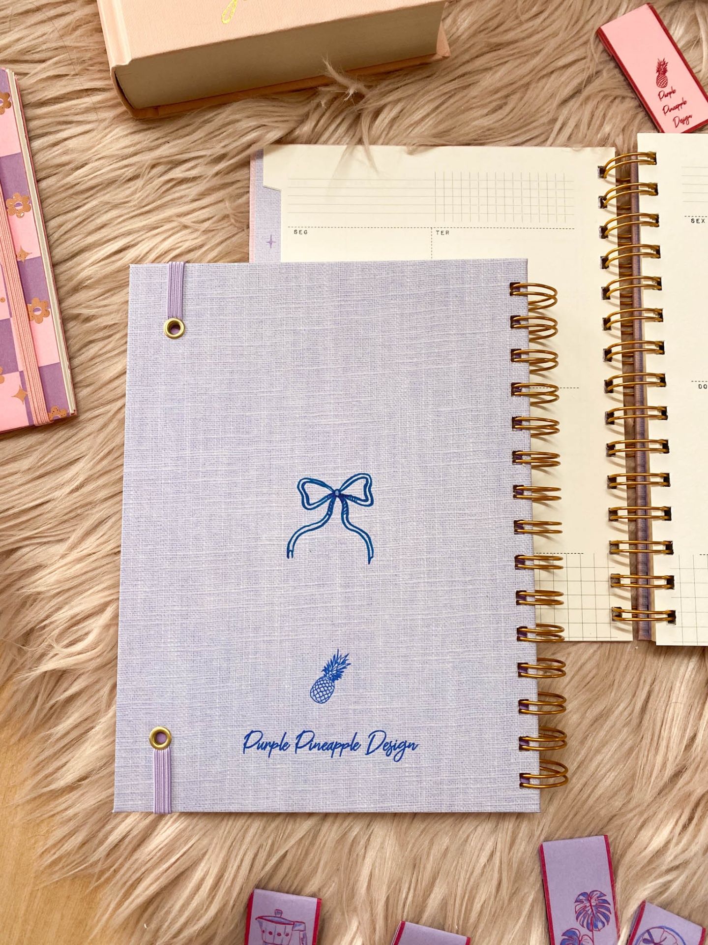 Caderno azul claro com laço azul, texto e abacaxi, encadernação dourada, sobre tapete peludo