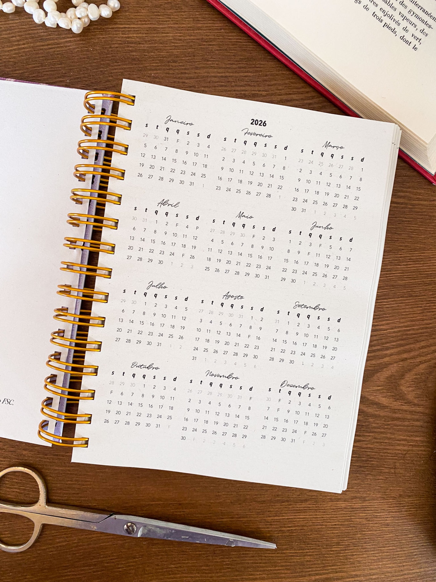 Agenda anual 2026 com espiral dourado e livro vermelho ao lado sobre mesa de madeira