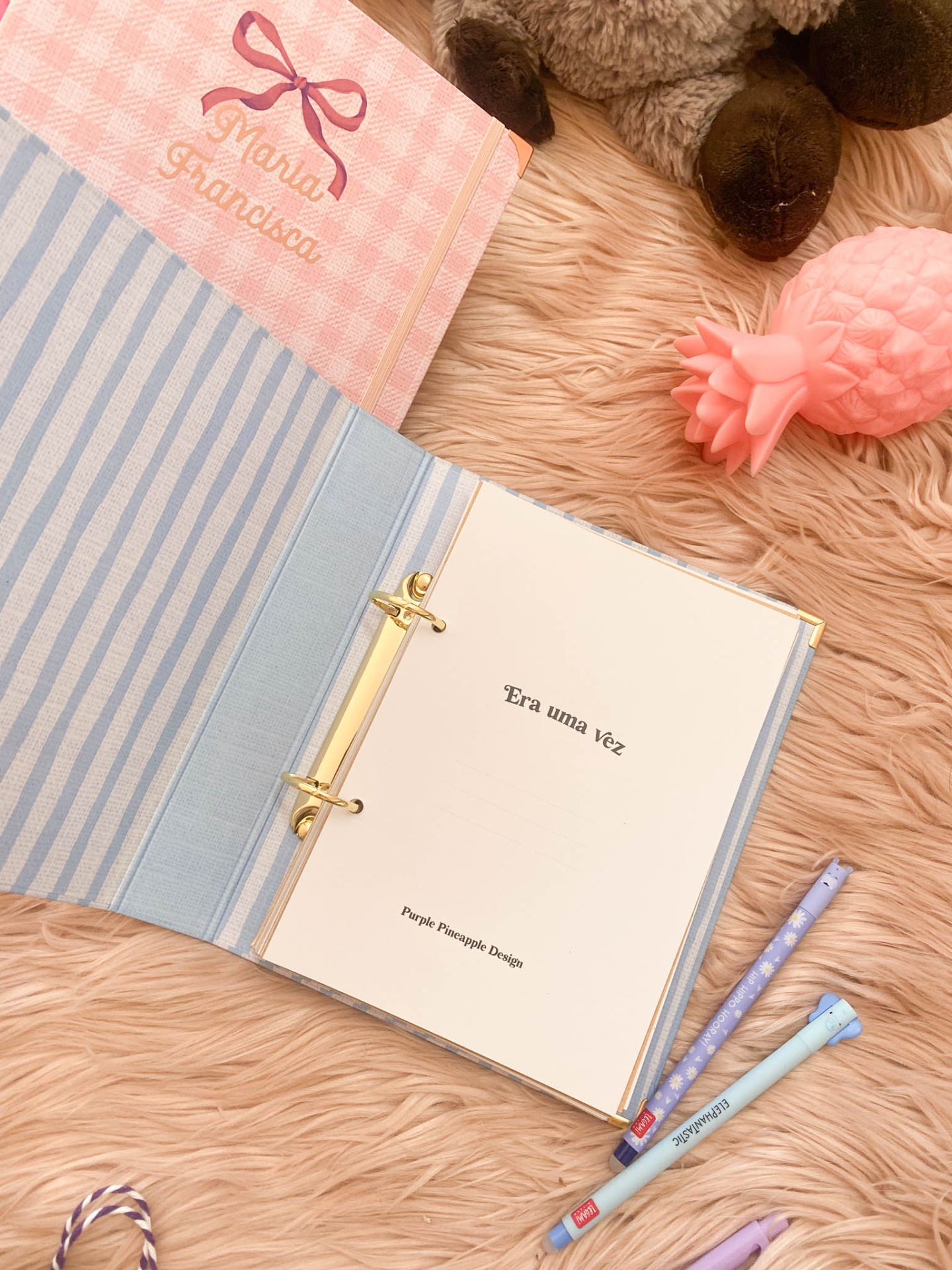 Caderno aberto e capa rosa com nome, peluche e brinquedo ananás em superfície cor de rosa