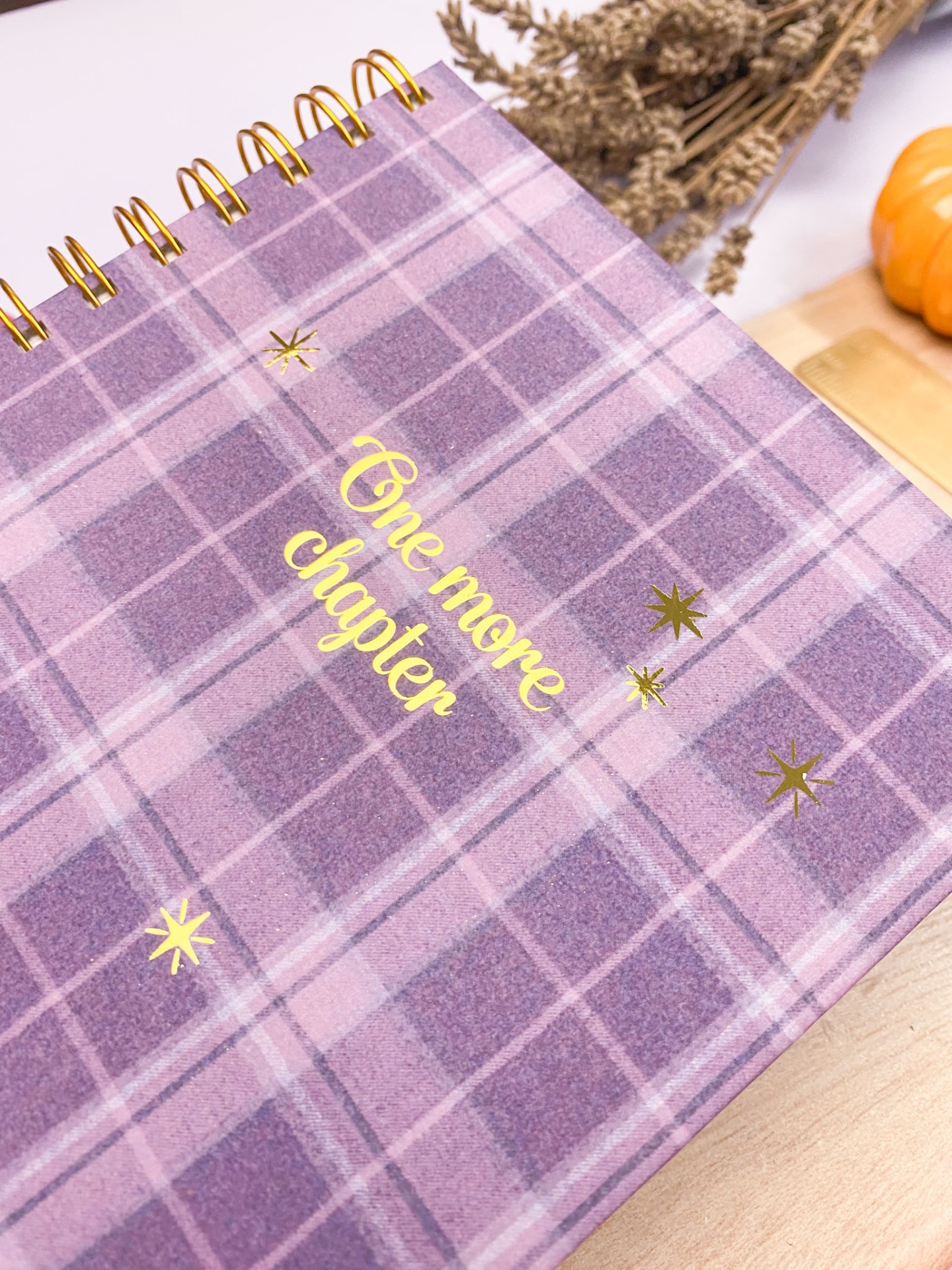 Caderno quadroxis lilás e roxo com texto e estrelas douradas em capa, espiral dourada, sobre mesa de madeira e decoração com plantas secas.