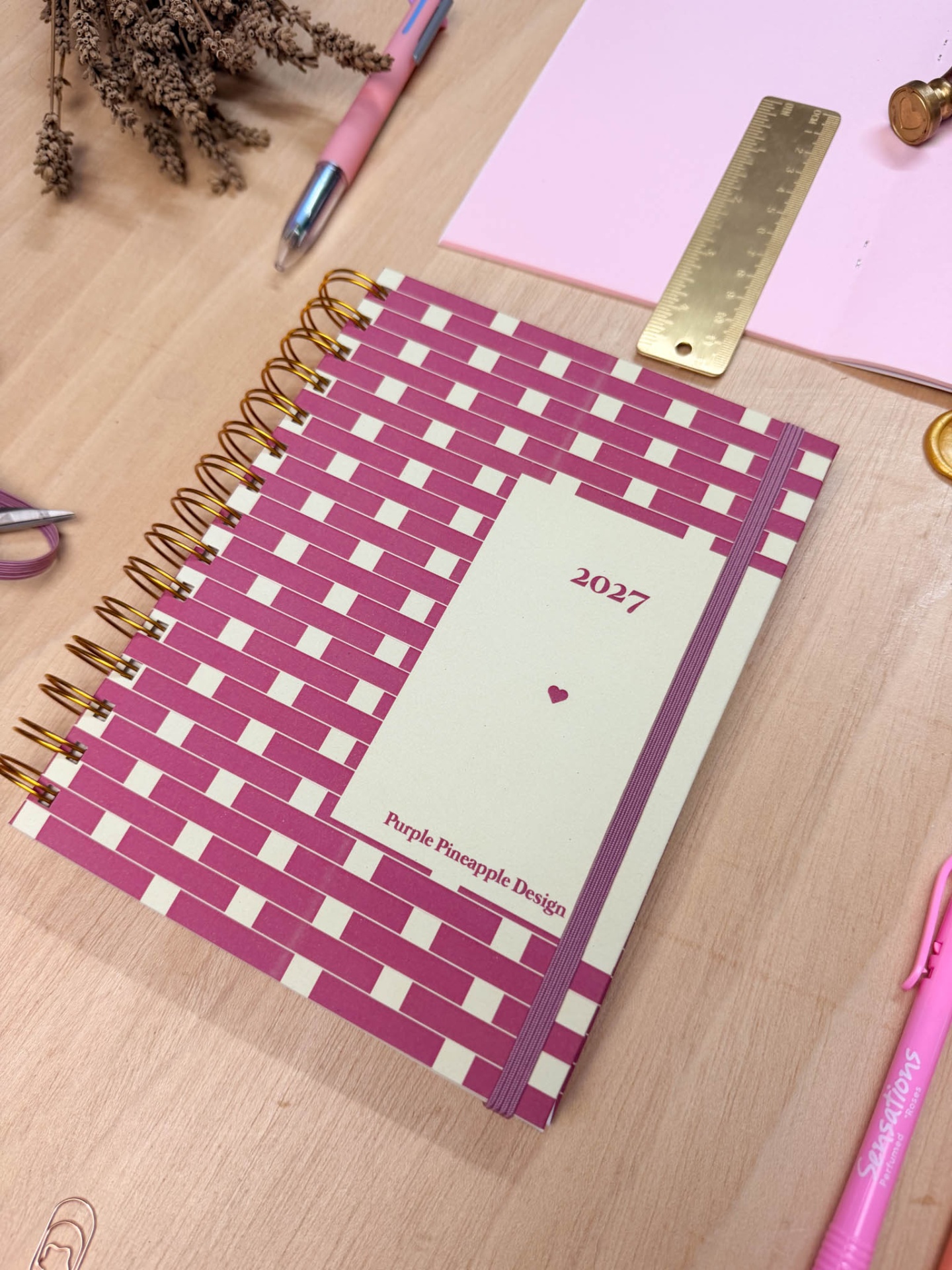 Caderno espiral rosa e branco com elástico, régua dourada, canetas rosa e flores secas em mesa de madeira clara