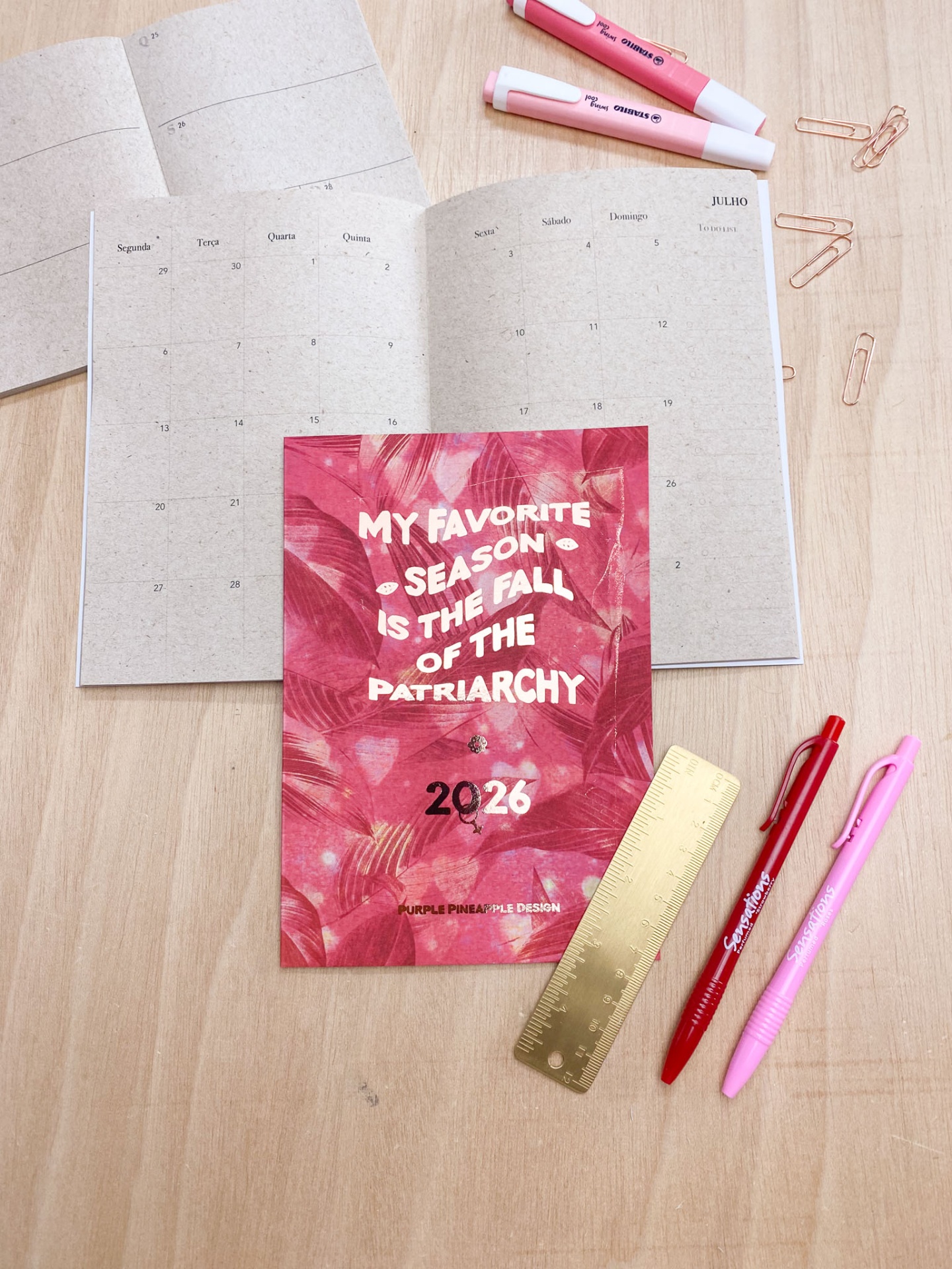 Planner 2026 com capa vermelha e padrão floral, texto branco, aberto sobre mesa de madeira com materiais de escrita