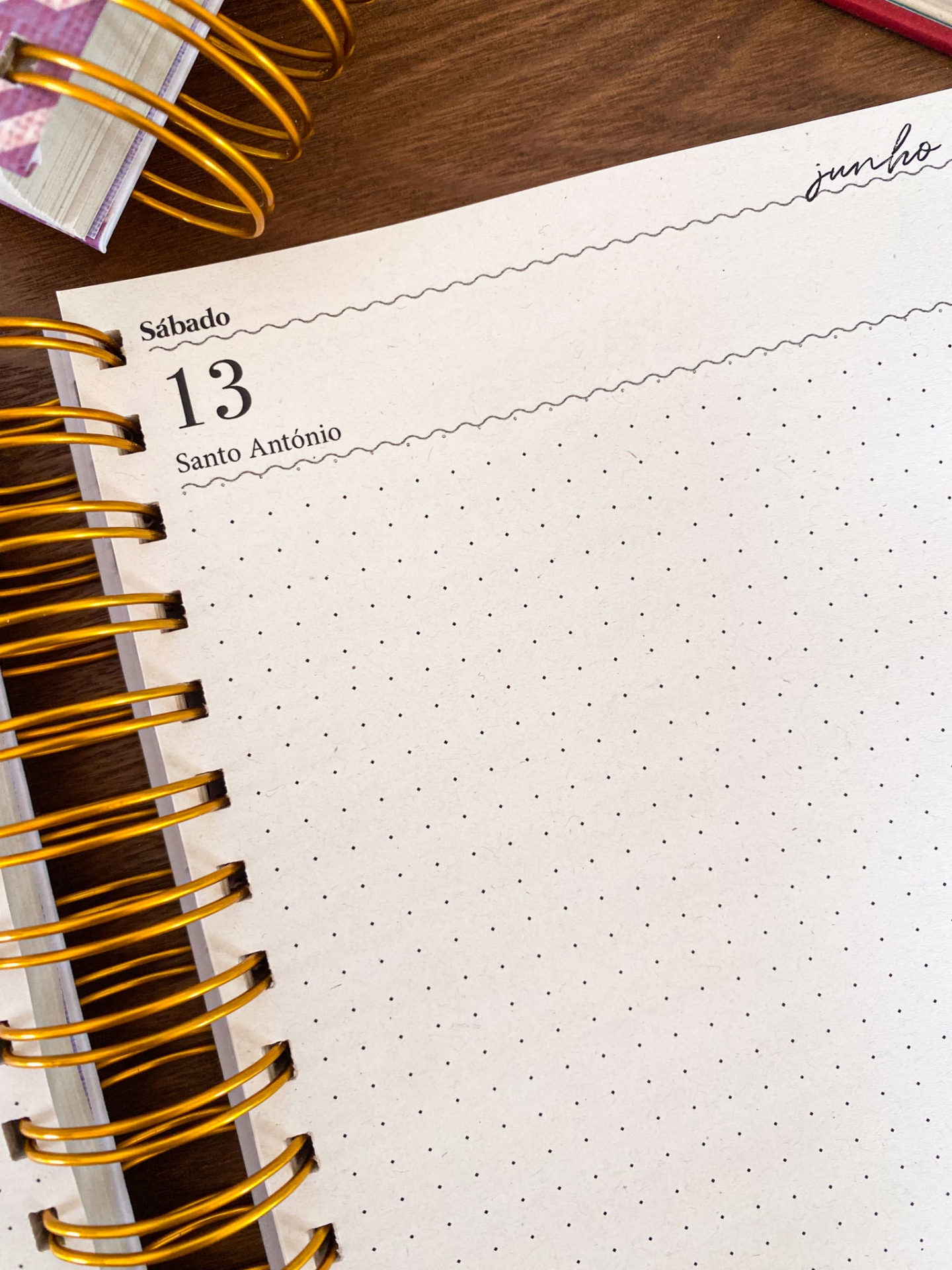 Página de agenda mensal pontilhada com espiral dourada em mesa de madeira.