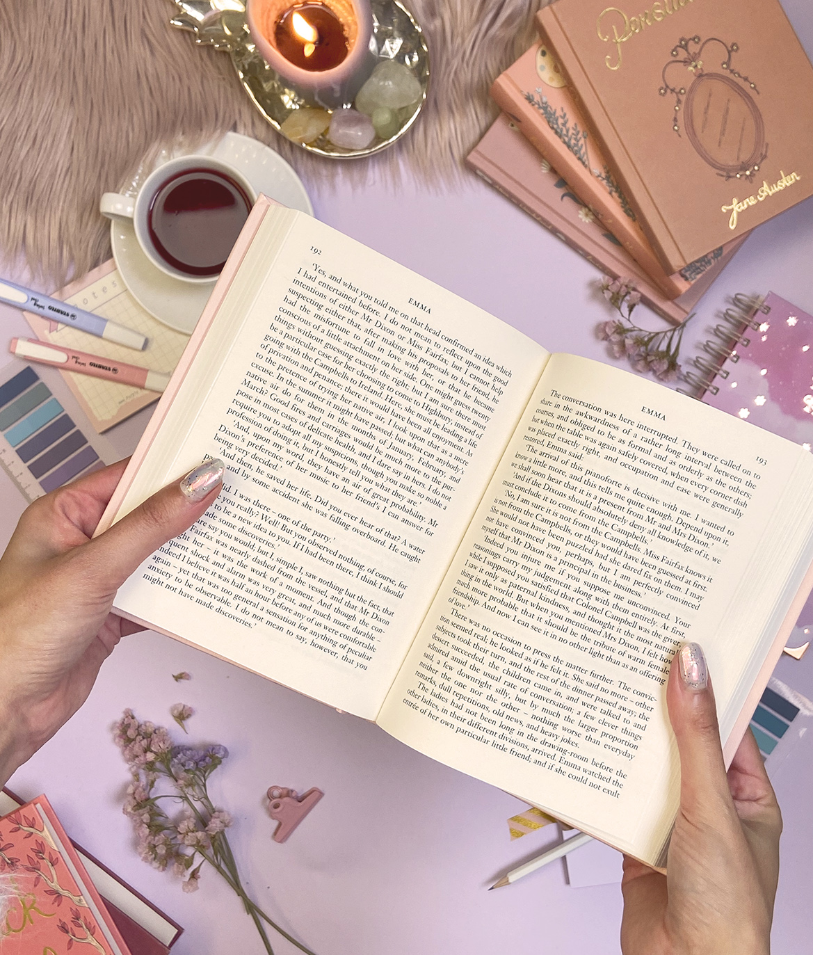 Livro aberto segurado por mãos, vela acesa, canetas, cadernos 'PARECER', chá, flores e itens de papelaria sobre mesa lilás.