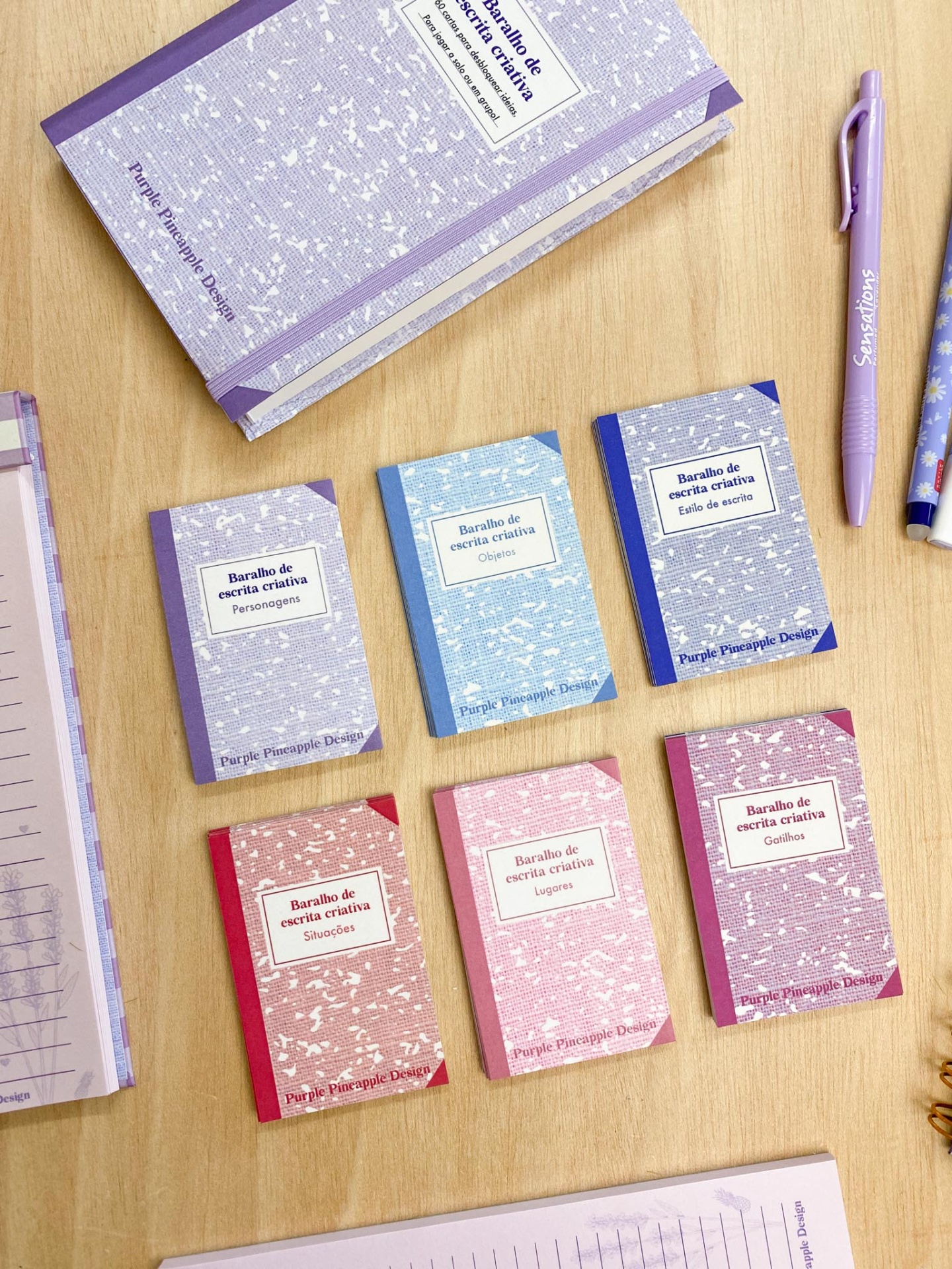 Seis blocos de notas pequenos coloridos com texto 'Baralho de escrita criativa' e canetas lilás numa mesa de madeira.