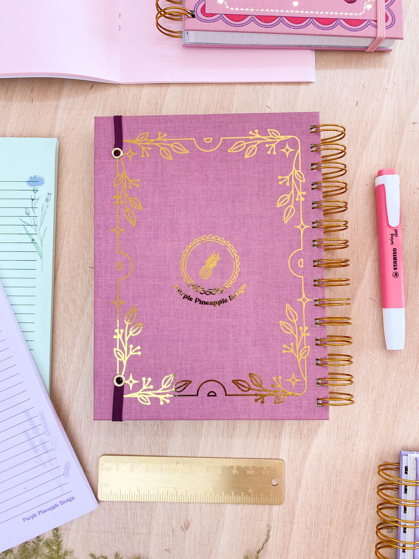 Caderno rosa com detalhes dourados e espiral, régua dourada, caneta rosa e outros cadernos numa mesa de madeira