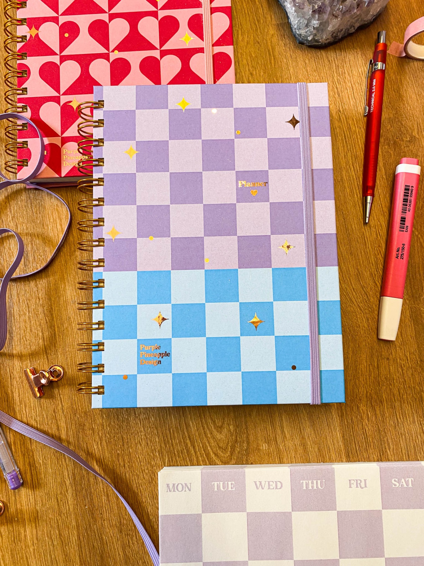 Diários com capa azul e lilás xadrez com espiral dourada e acessórios de escrita numa mesa de madeira
