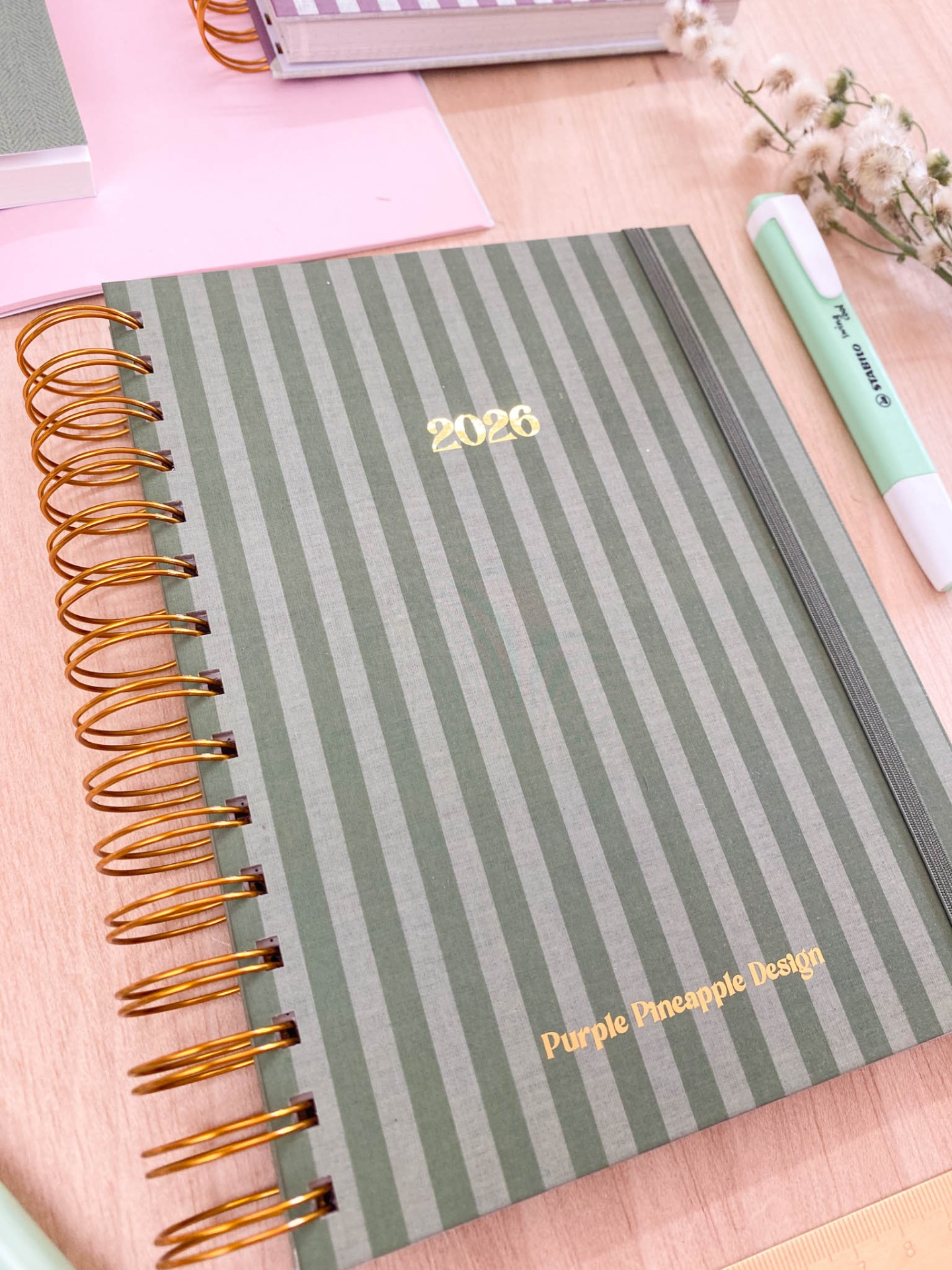 Caderno verde às riscas com espiral dourada e texto '2026' e 'Purple Pineapple Design'