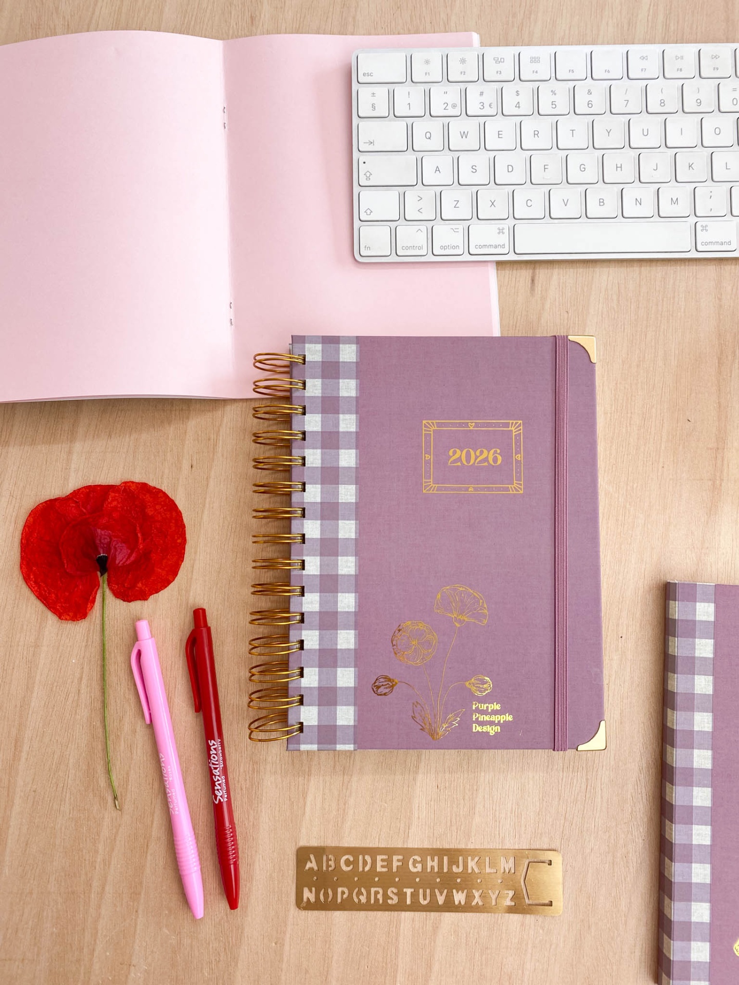Agenda 2026 roxa com detalhes dourados, canetas, flor vermelha, teclado branco, capa rosa aberta e régua alfabética sobre mesa de madeira