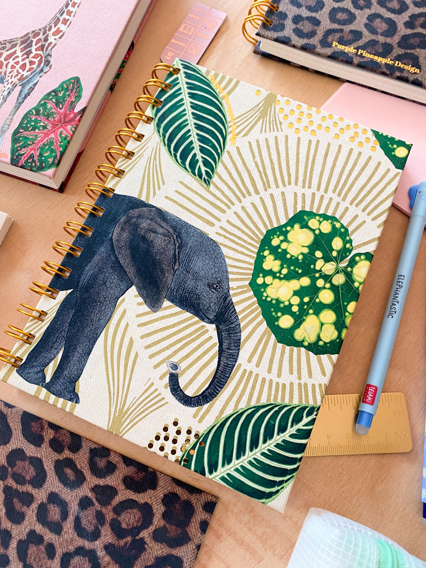 Caderno espiral com capa ilustrada de elefante e folhas verdes e douradas em mesa com outros itens de papelaria